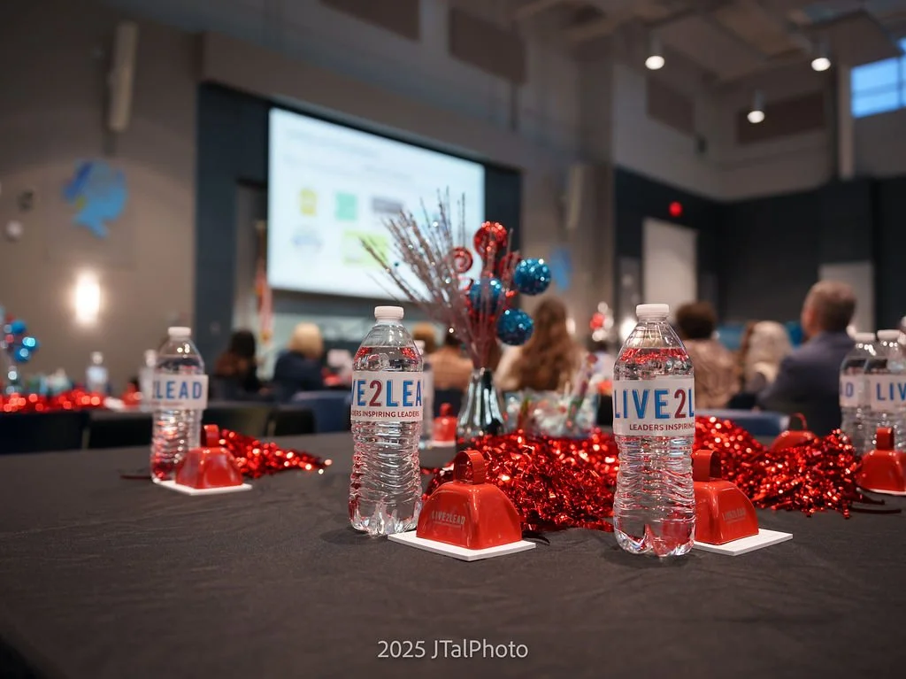 Conference room with tables decorated for the holidays, featuring water bottles labeled 'LIVE2LEAD', red tinsel, and a centerpiece with blue and red ornaments. Attendees are seated facing a large screen at the front.
