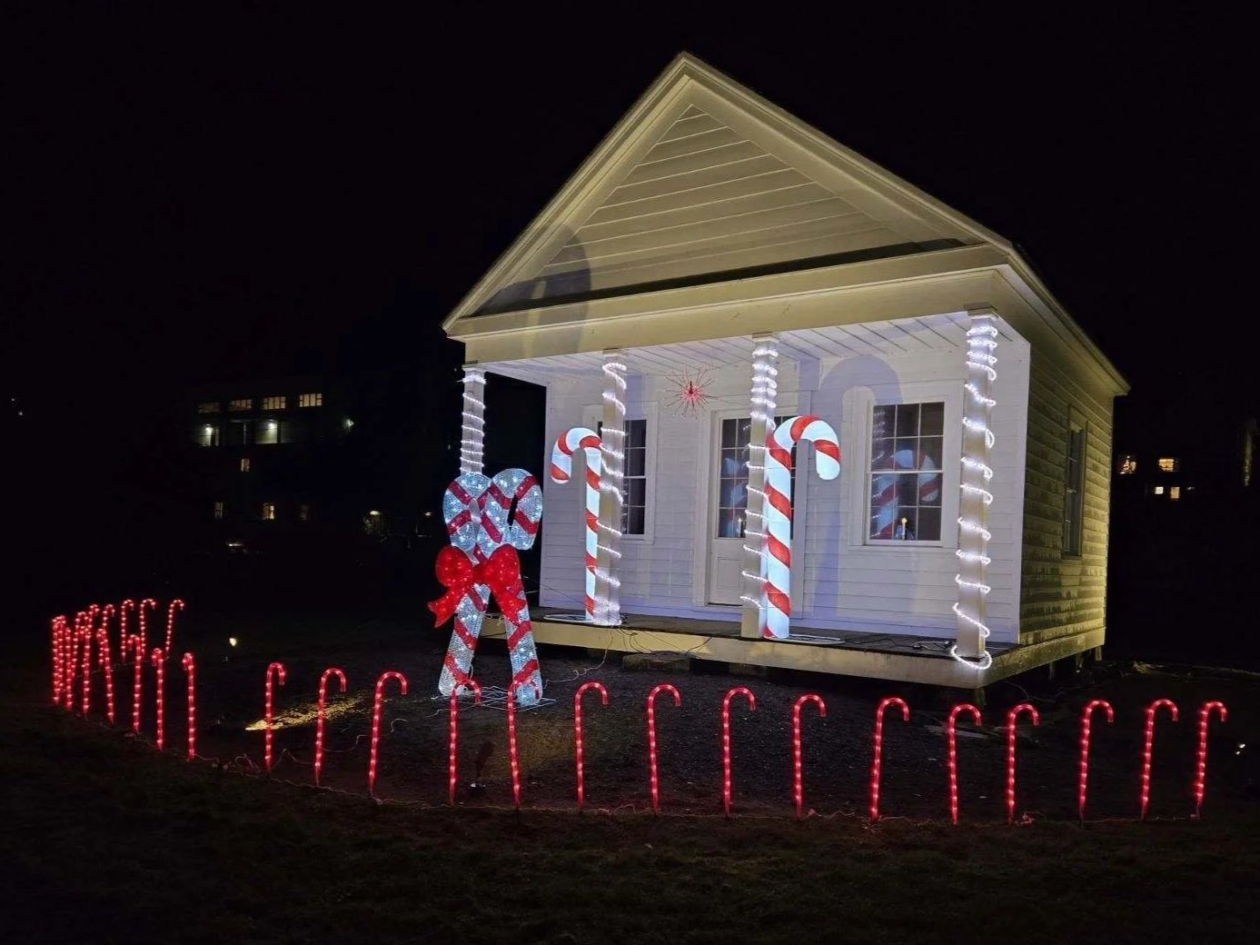 Small house decorated with Christmas lights and large candy cane props on porch, surrounded by red light fence, during night.
