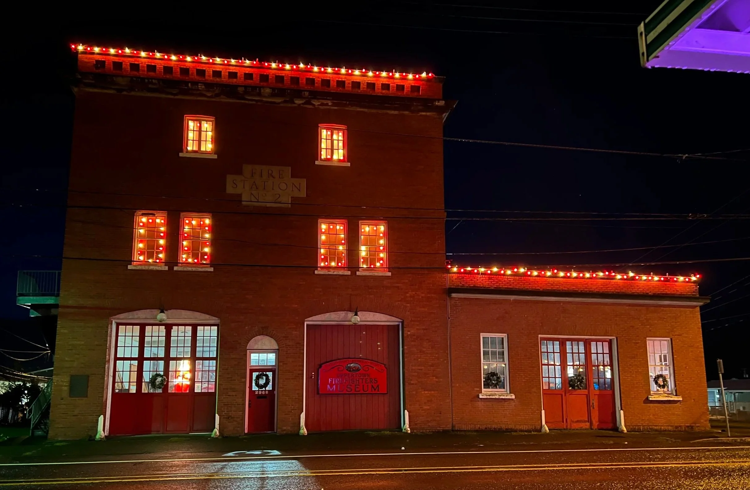 Night view of a historic brick fire station building decorated with red lights and Christmas wreaths, featuring a sign that reads 'Fire Station No. 2' and a Museum sign on the door.