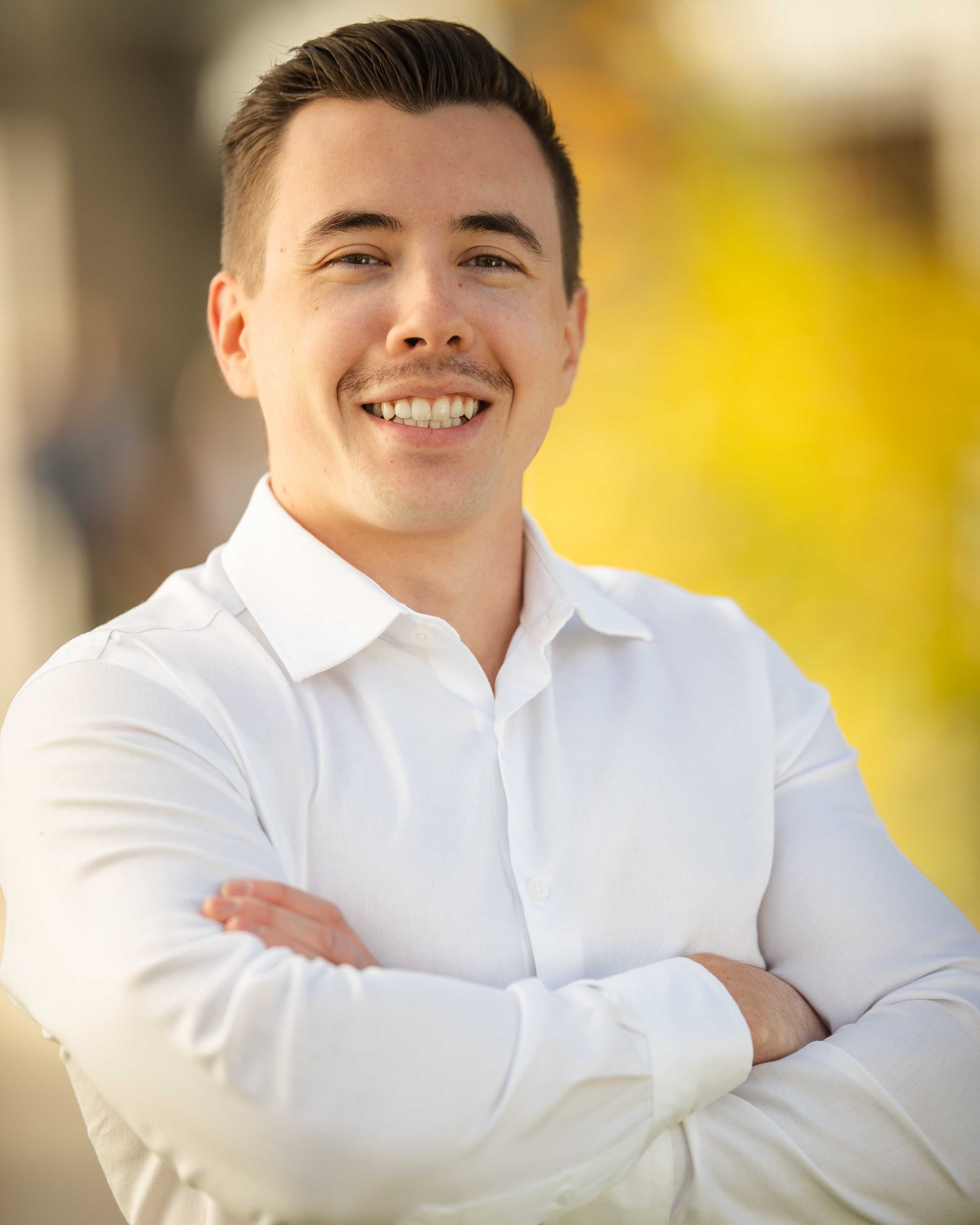 A young man smiling with arms crossed, wearing a white button-up shirt outdoors with a blurred background of trees and yellow foliage.