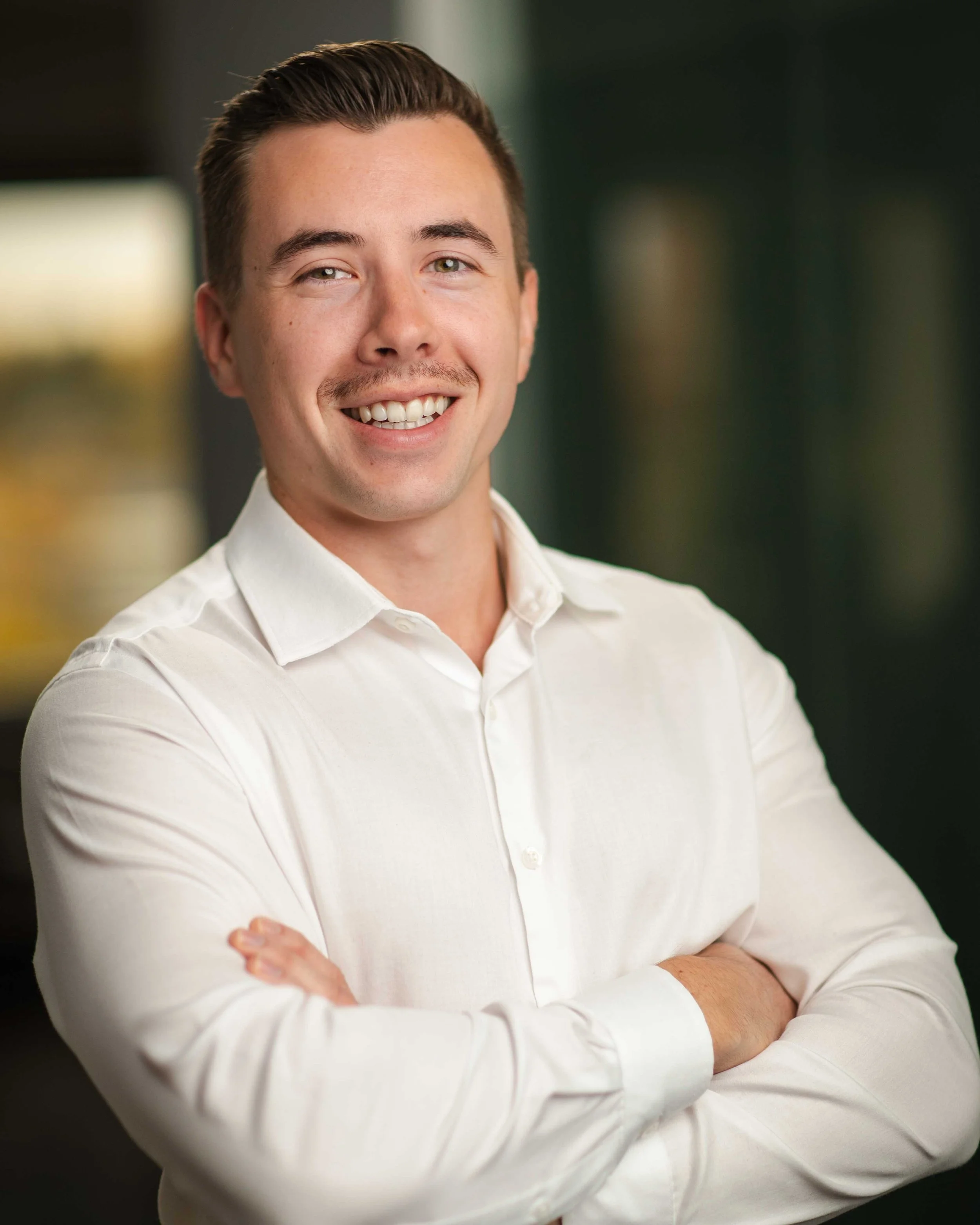 A young man with short dark hair, wearing a white button-up shirt, smiling and crossing his arms, standing indoors with a blurred background.