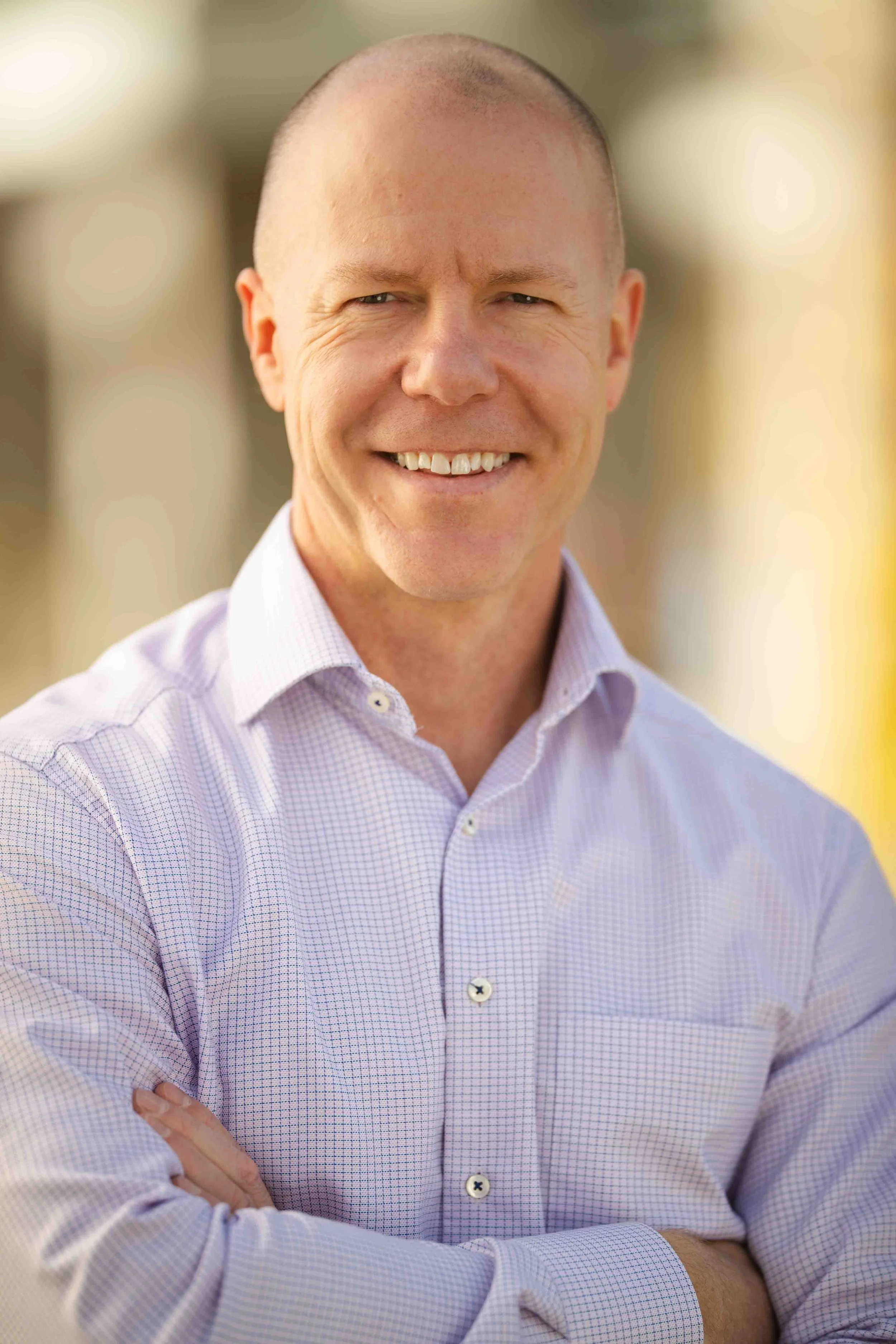 A smiling man with a shaved head wearing a light purple button-up shirt with crossed arms, standing outdoors with a blurred background.