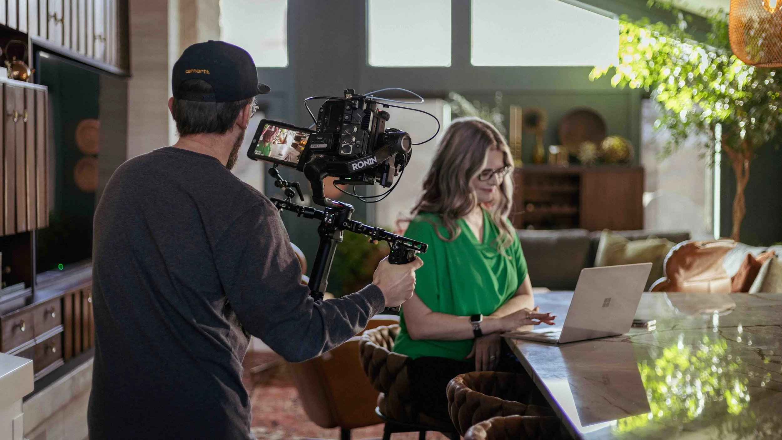A person with a camera filming a woman sitting at a table working on a laptop in a well-lit living room.