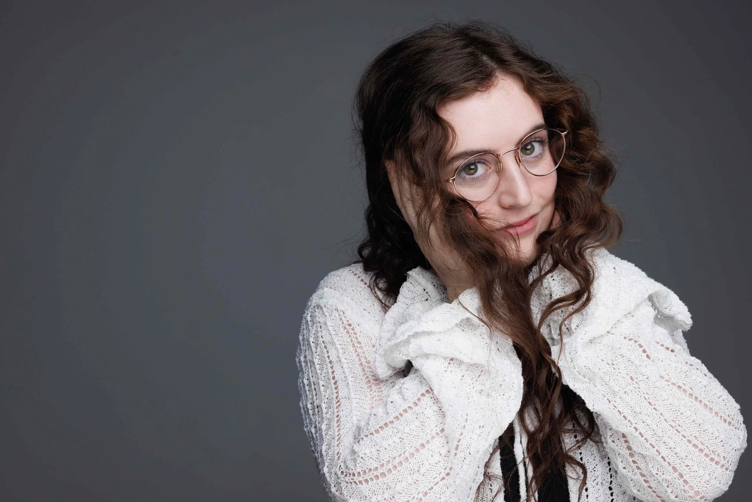 Young woman with curly brown hair and glasses, wearing a white lace top, posing against a gray background.