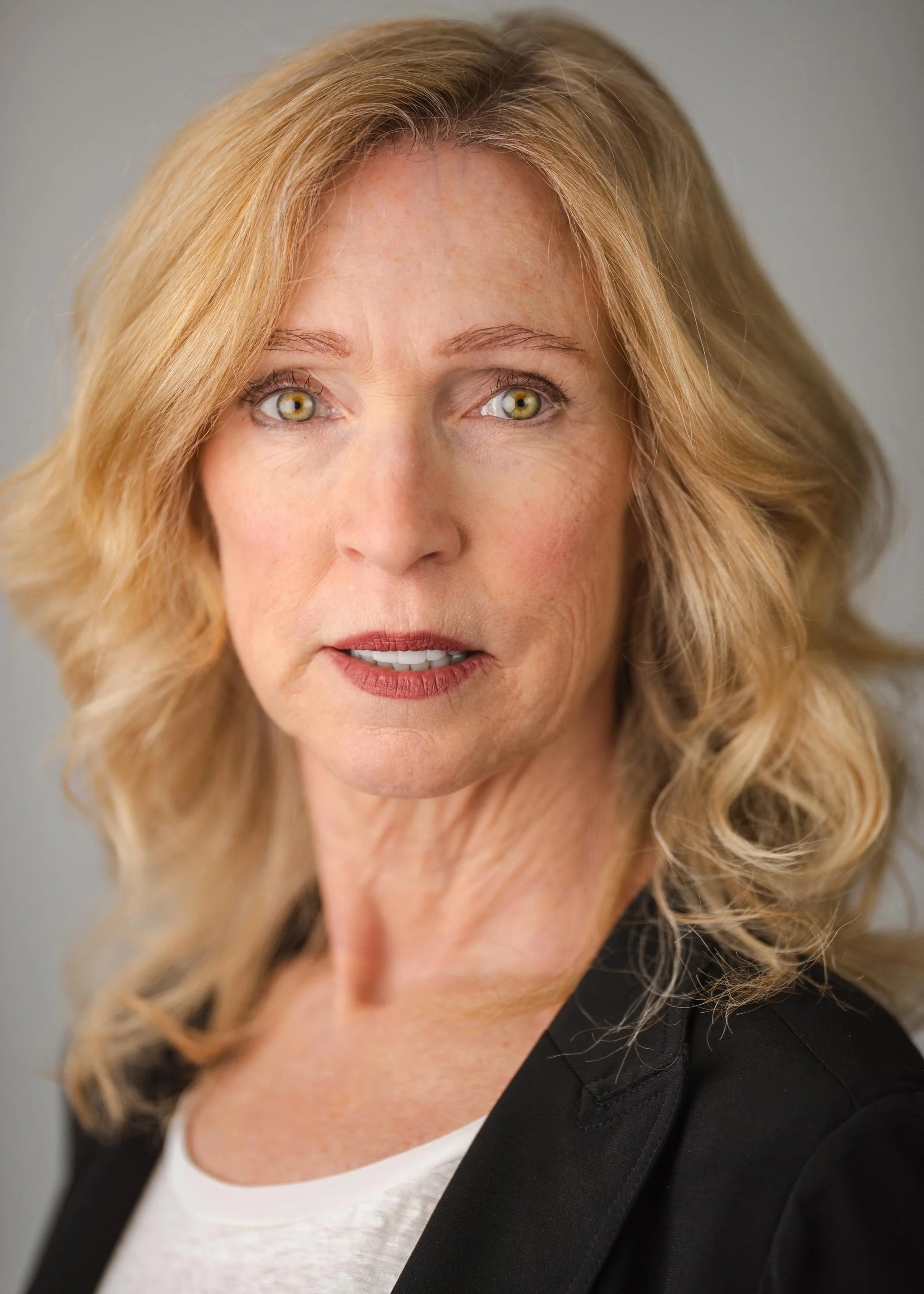 Close-up portrait of a middle-aged woman with blonde wavy hair, light makeup, and wearing a black blazer over a white top, looking directly at the camera with a neutral expression.