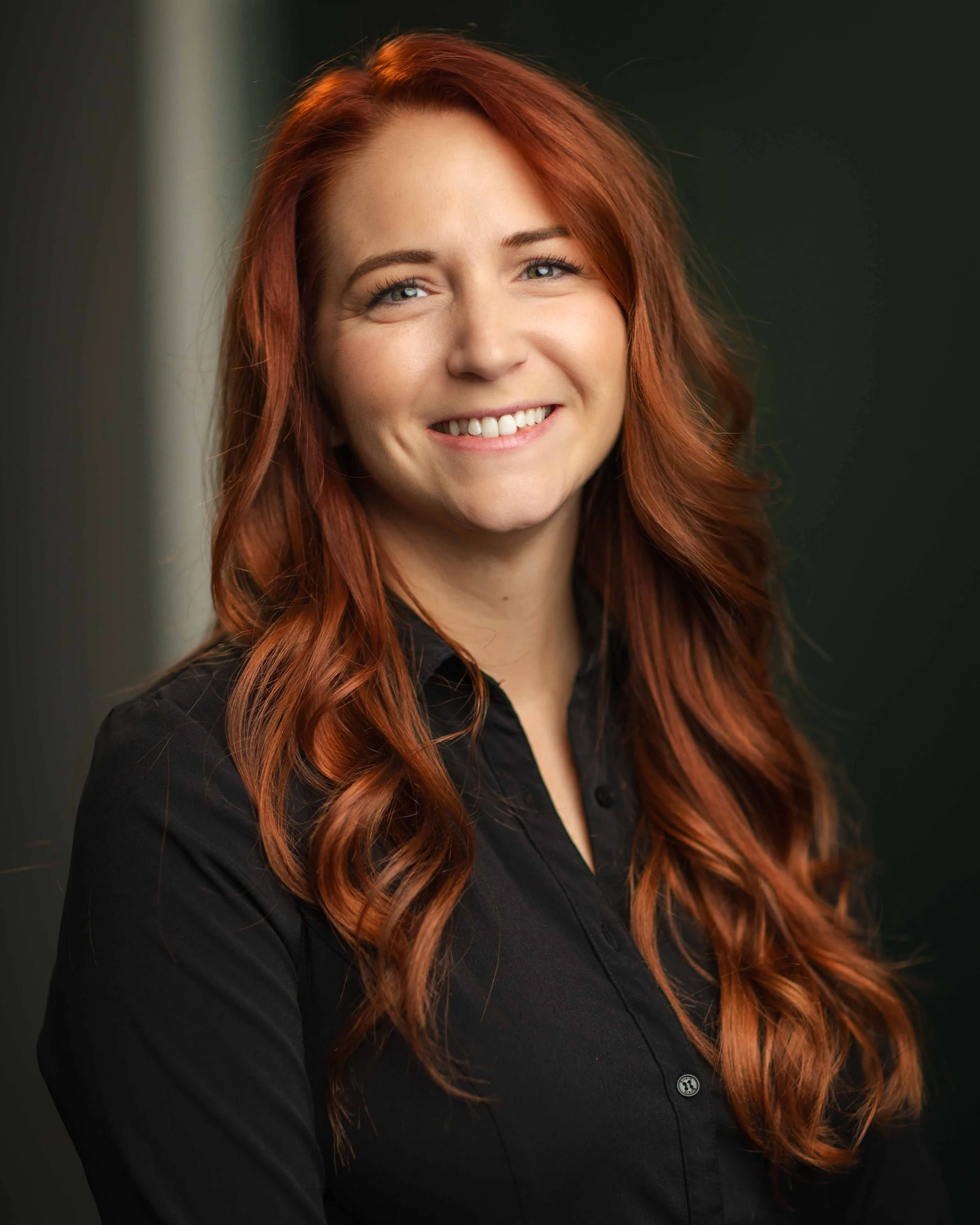 A woman with long, wavy red hair smiling, wearing a black button-up shirt, standing against a dark background.