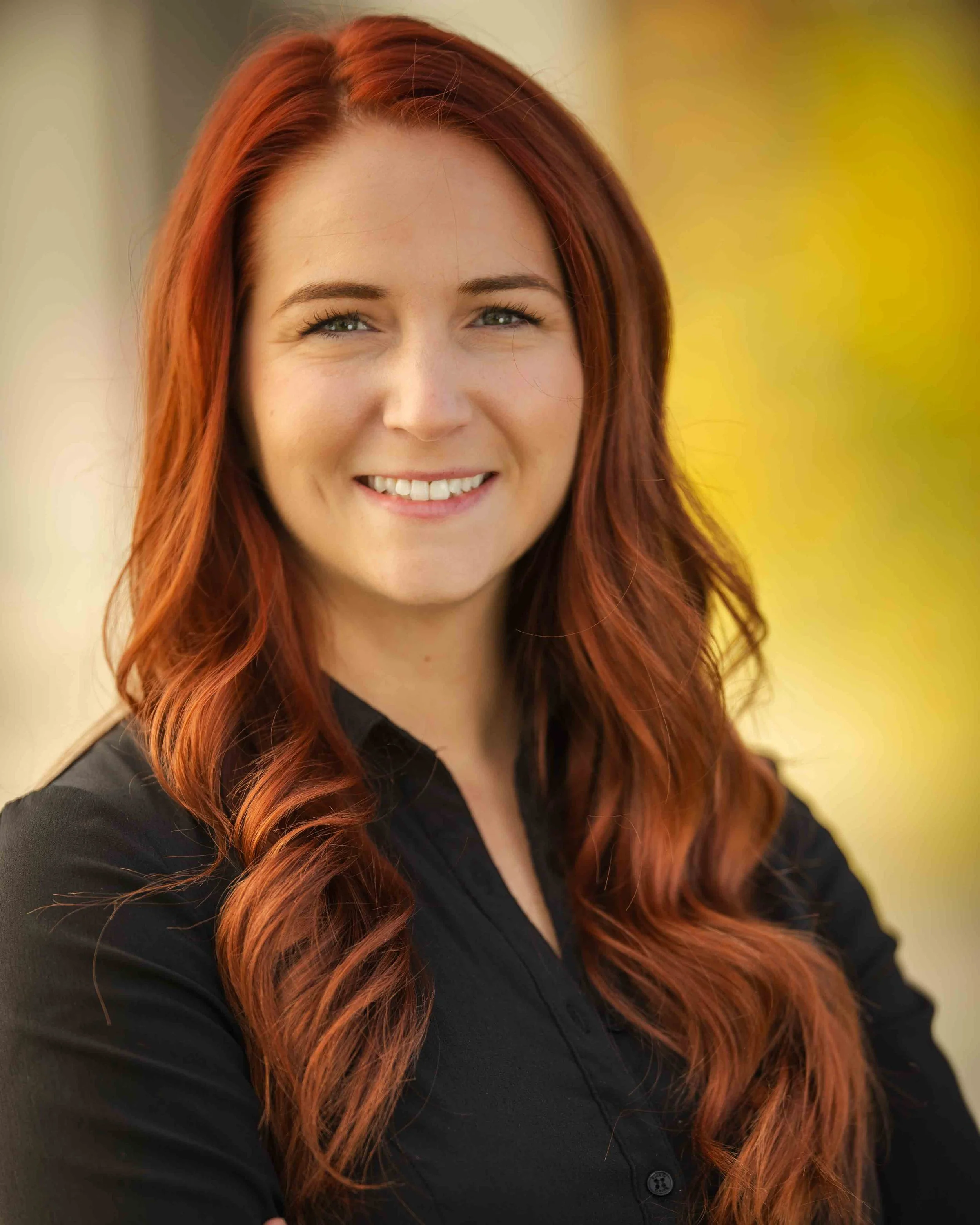 A woman with long red hair smiling outdoors with a yellow blurred background.