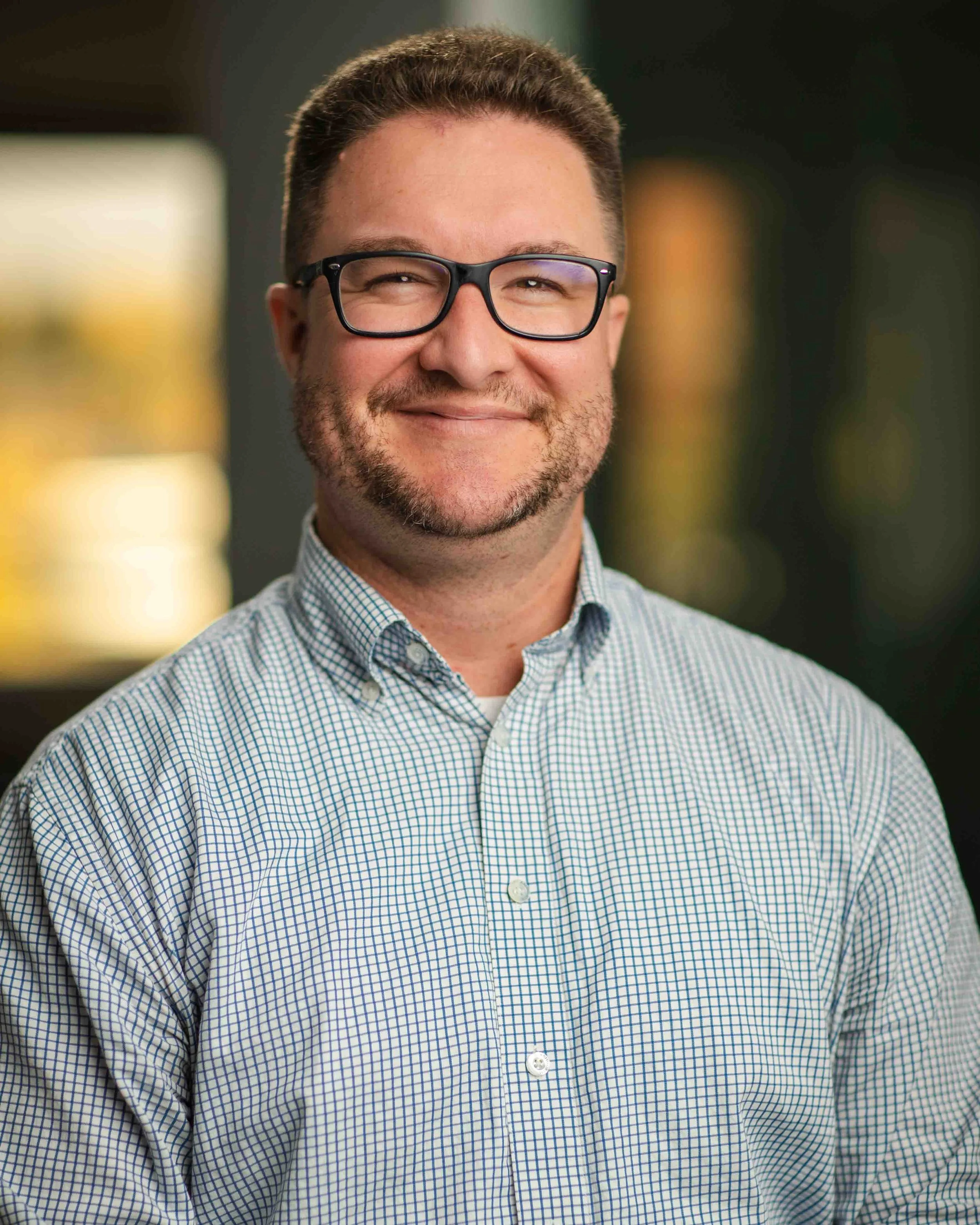 A smiling man with glasses, short brown hair, and a beard, wearing a light blue checkered shirt, standing indoors with a blurred background.