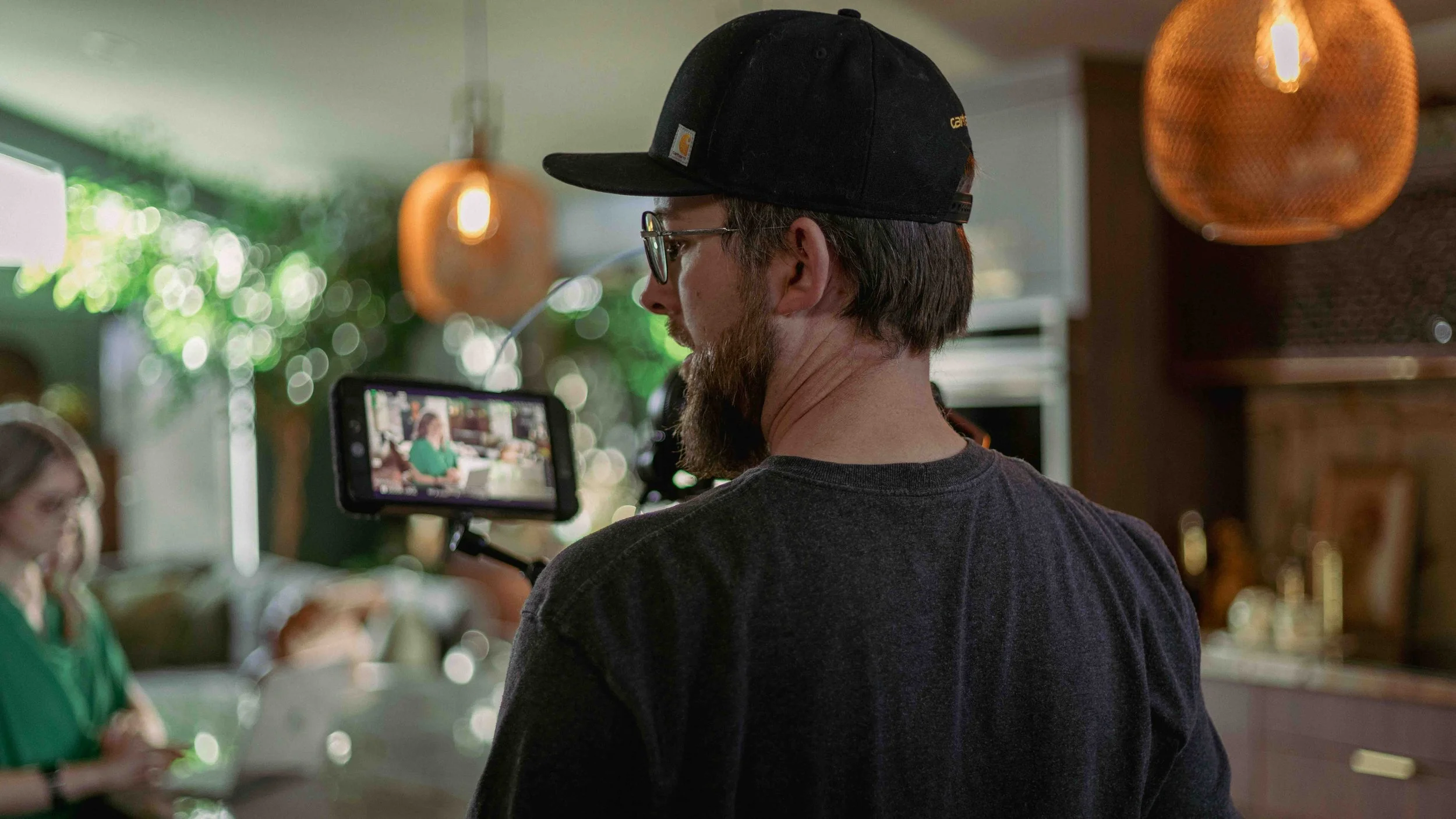 A man wearing glasses, a black cap, and a dark t-shirt standing in a kitchen, filming with a camera mounted on a tripod. In the background, a woman in a green shirt is sitting at a table using a mobile device.