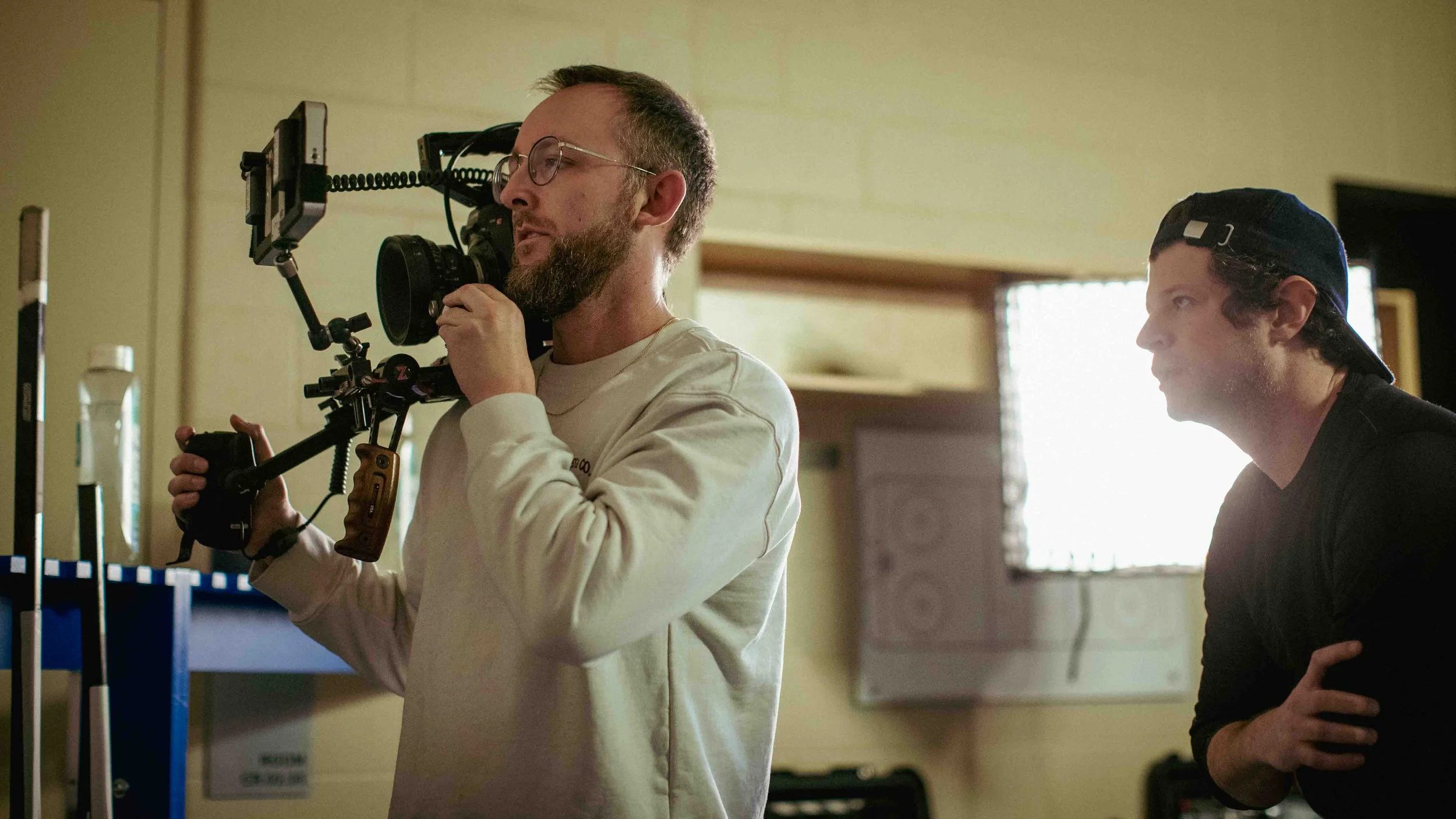 Two men on a film set, one holding a camera rig and wearing glasses, the other with a cap, both indoors near a window with blinds.