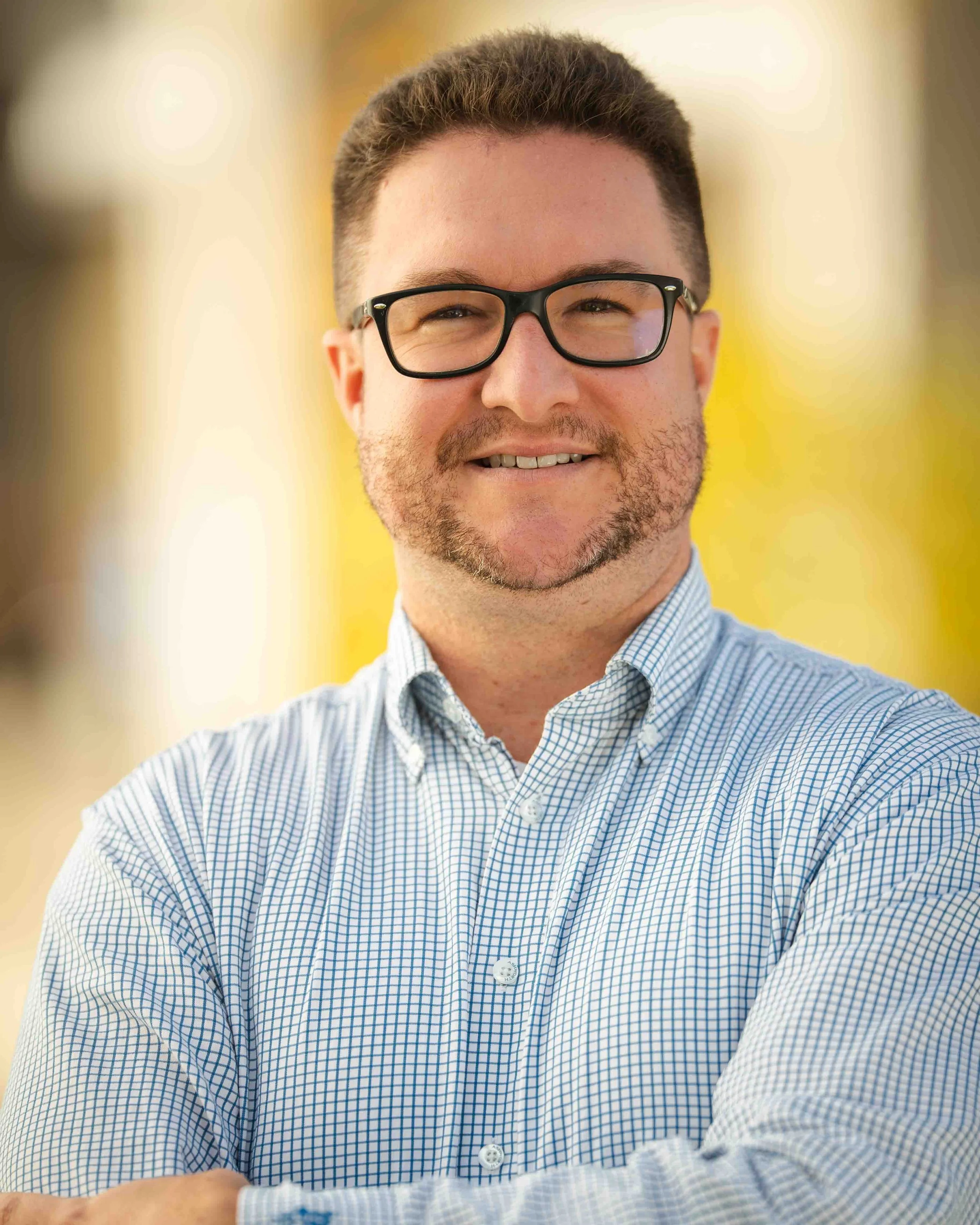 A man with short hair, glasses, and a beard is smiling and crossing his arms. He is wearing a light blue patterned button-up shirt, and the background is blurred with warm tones.