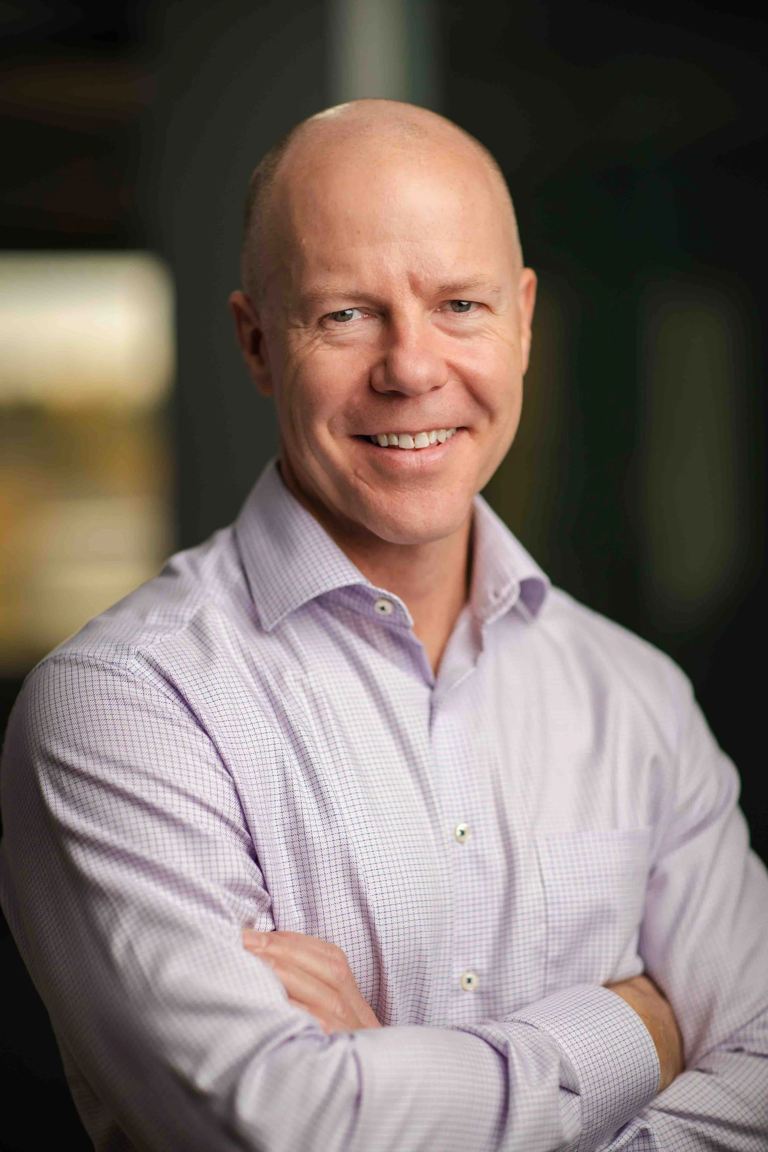 A smiling, bald man wearing a light purple checkered dress shirt with arms crossed, standing indoors with a blurred background.
