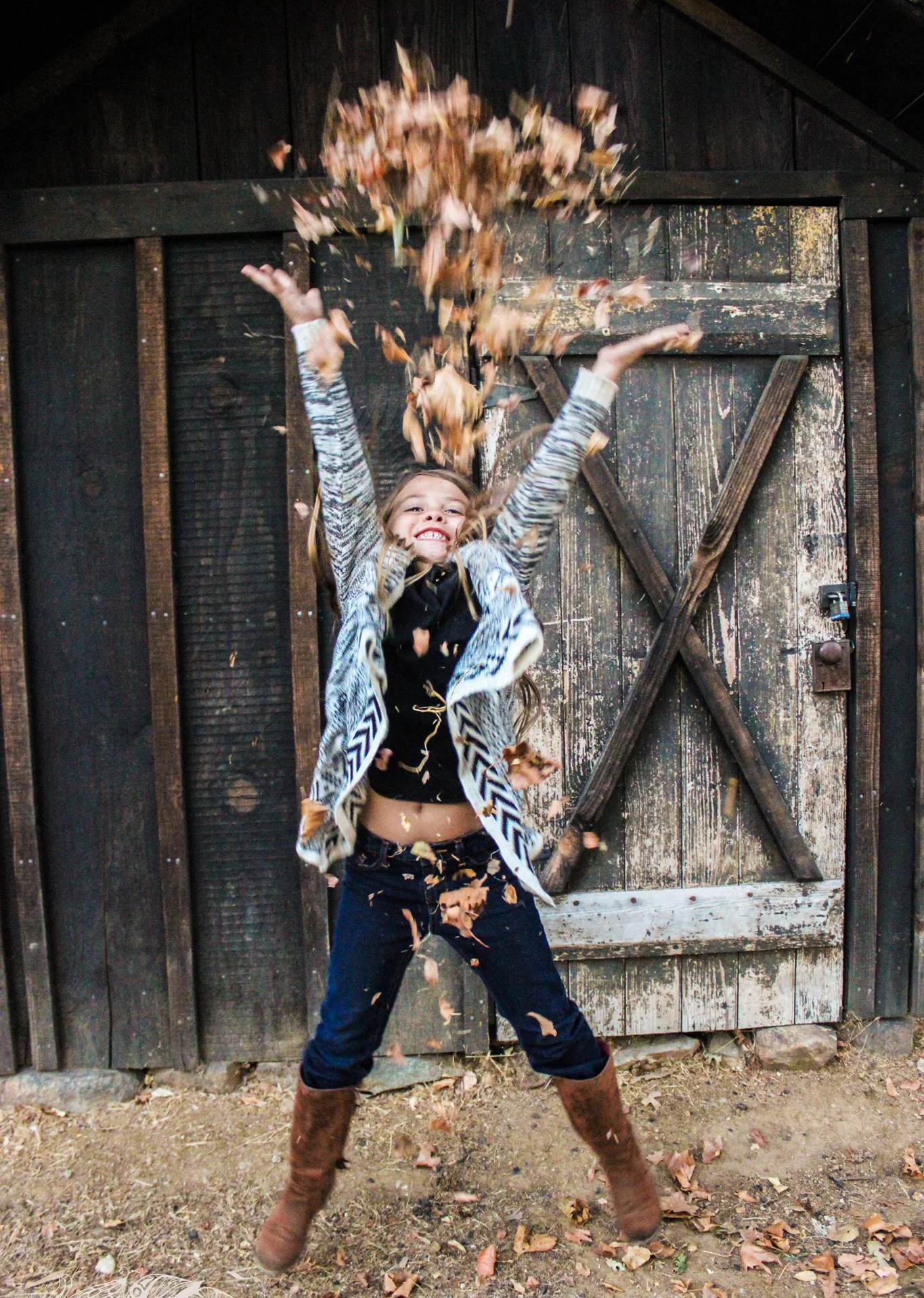 A girl in a patterned jacket and black top throws leaves in the air while standing in front of a weathered wooden barn door, smiling with joy.