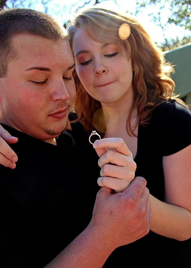 A woman appears to be proposing to a man, holding a ring in her hand as they both look at it.