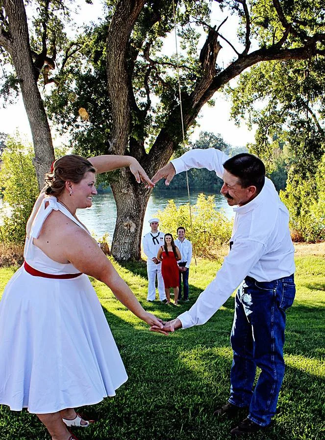 A couple holding hands and dancing outdoors near a tree, with a pond and three people standing in the background during daytime.