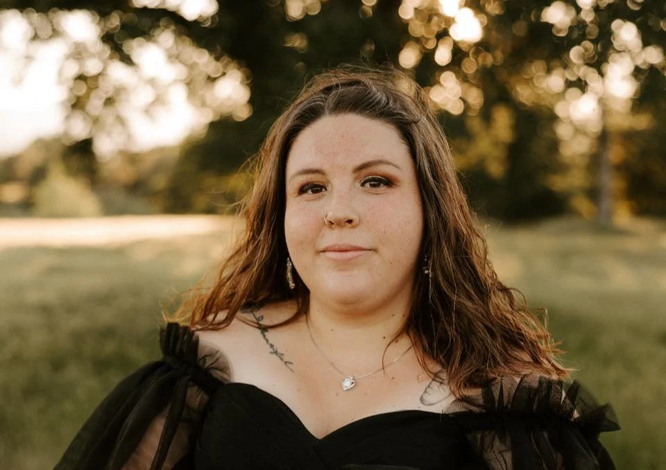 A woman with long wooden brown wavy hair wearing a black off-shoulder dress is outdoors during sunset, standing in a grassy area with trees in the background.