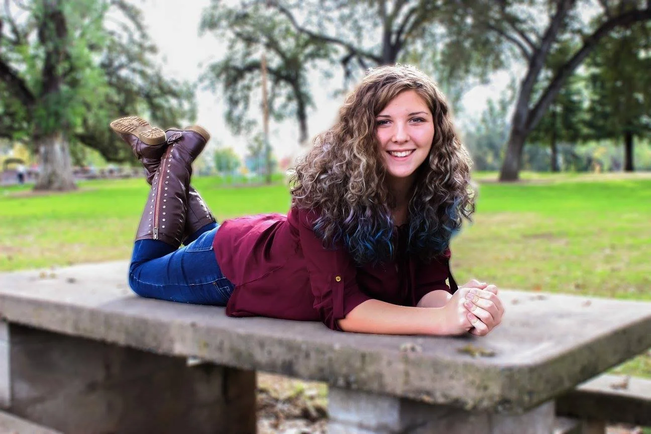 A young woman with curly hair lying on her stomach on a concrete park bench, smiling at the camera.