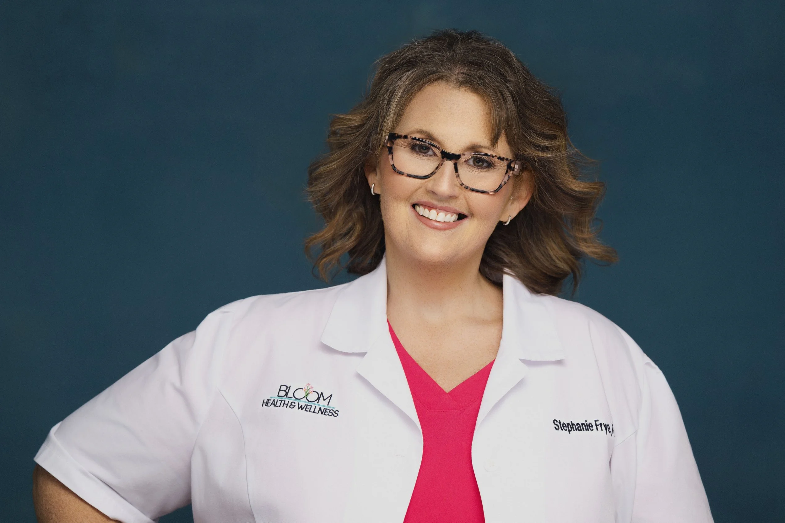 A woman with curly brown hair, glasses, and earrings, wearing a white medical coat over pink scrubs, standing against a dark background.