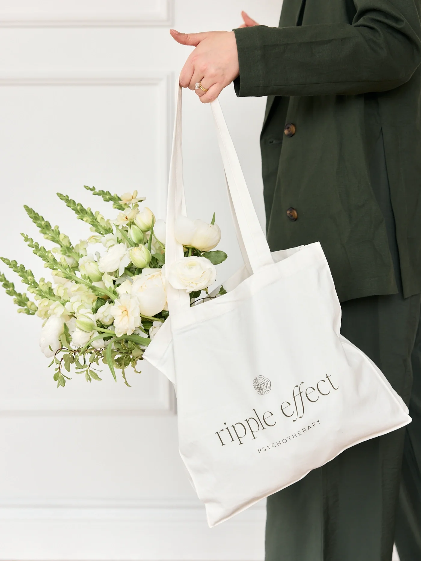 Person in dark green jacket holding a white tote bag with black text that says 'ripple effect psychotherapy'. The person is also holding a bouquet of white and green flowers.