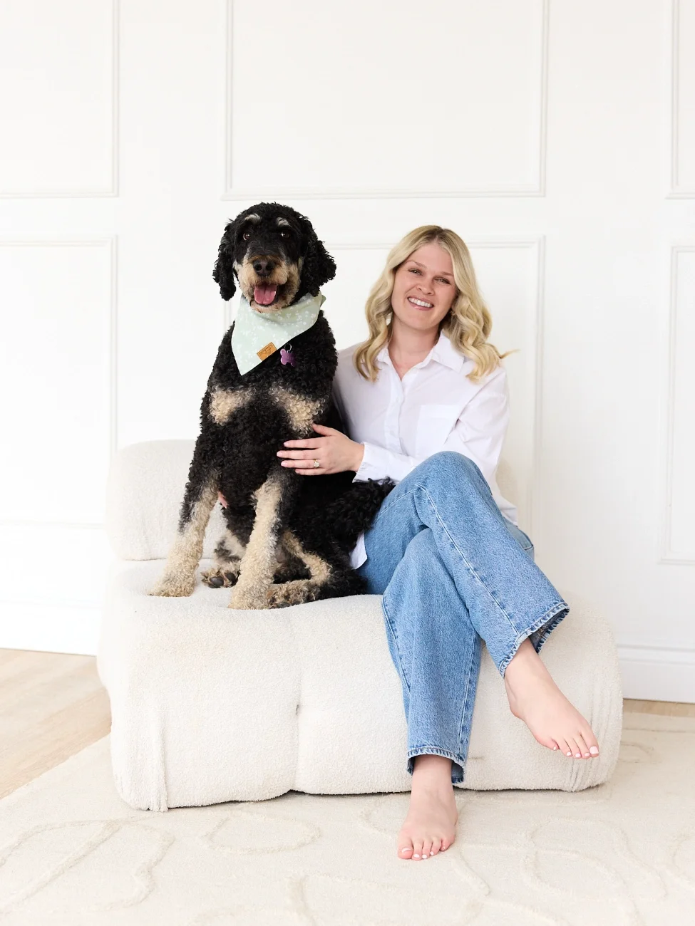 A woman sitting on a white couch, smiling, with a large black and tan dog sitting beside her. The dog wears a light-colored bandana and has its tongue out.