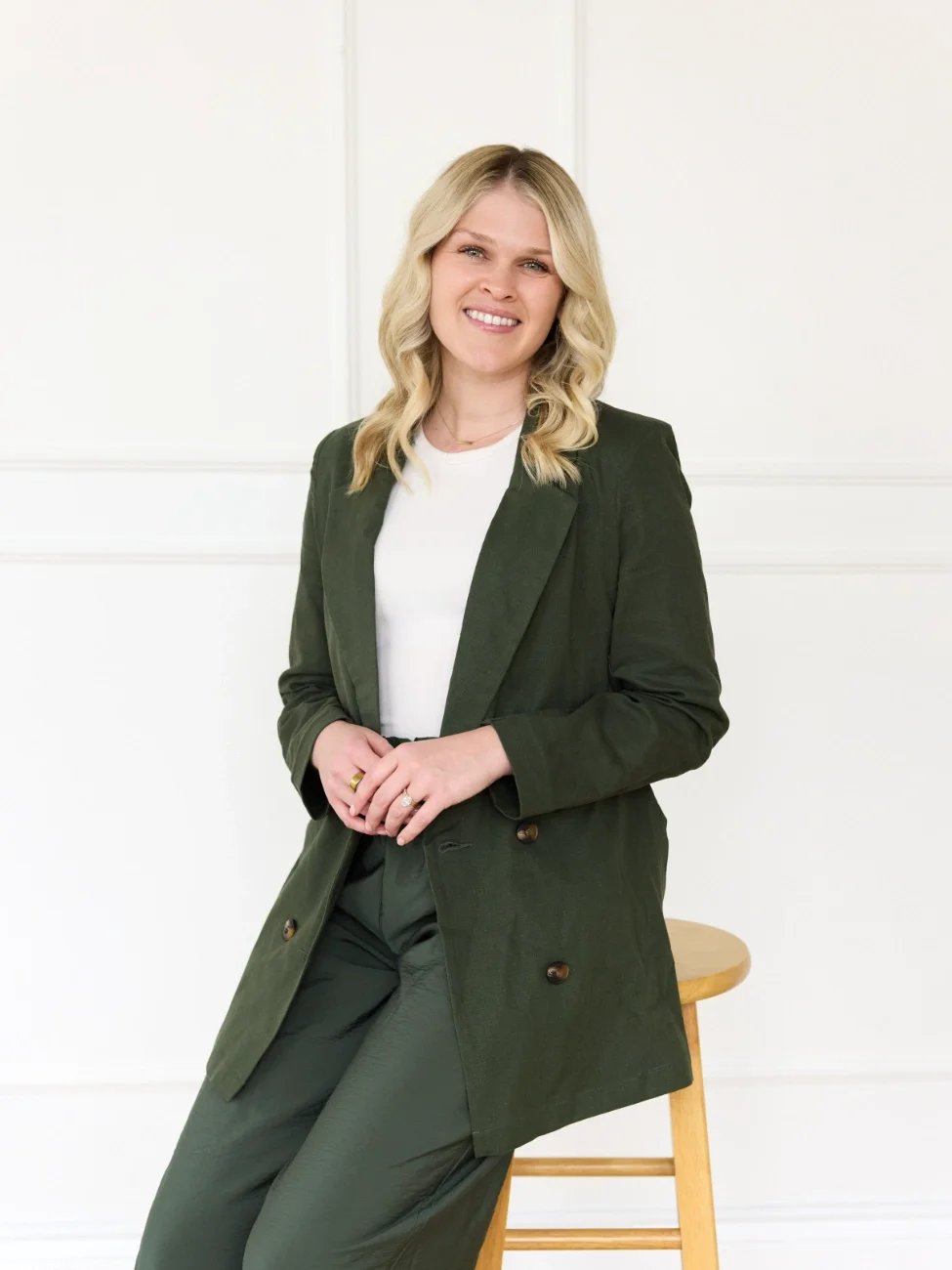A smiling woman with blonde hair, wearing a dark green blazer and pants, sitting on a wooden stool against a white wall.