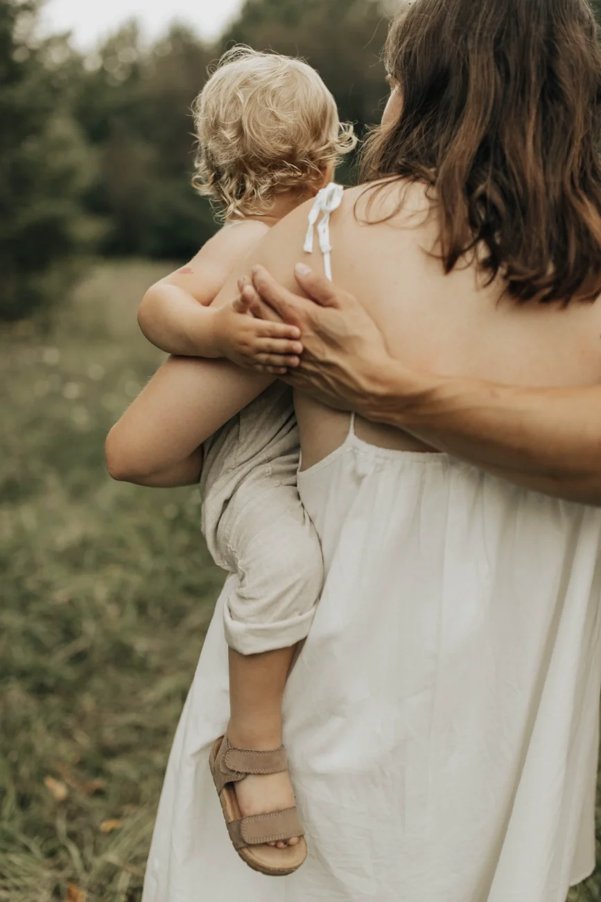 An adult woman holding a young child outside by a body of water. The woman is supporting the child with her arm and hand, and the child, who has curly blond hair, is looking away from the camera. The scene appears to be in a natural outdoor setting with trees in the background.