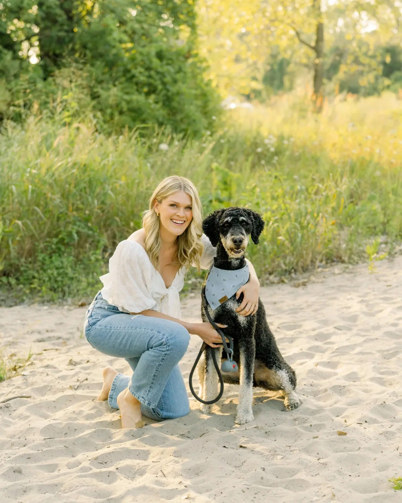 A woman with blonde hair, wearing a white blouse and jeans, kneels on the sand next to a black and white dog wearing a blue bandana and harness, on a sandy outdoor path with green foliage and trees in the background during daytime.