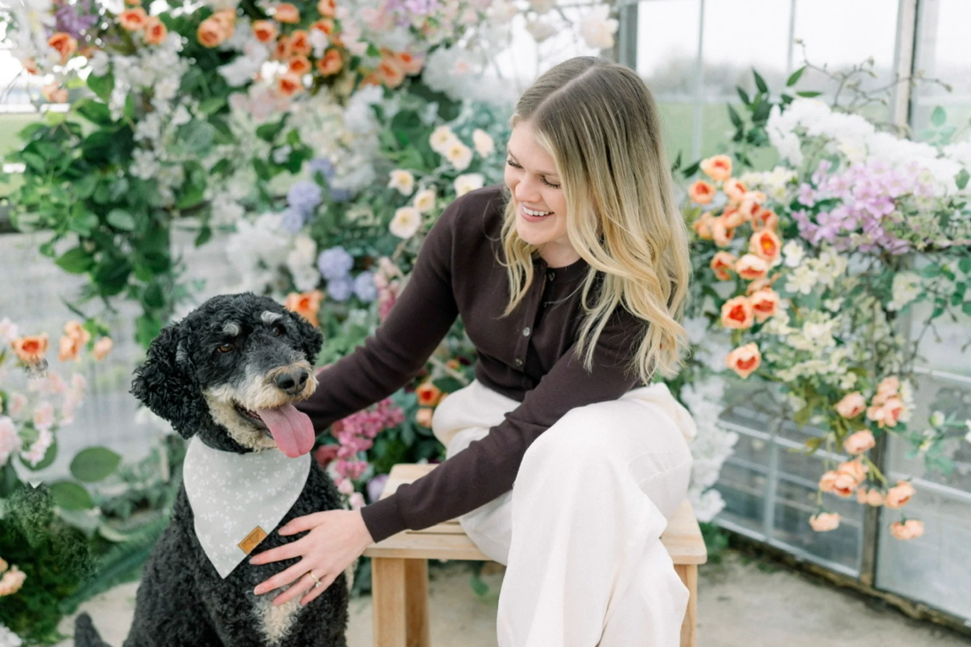 A woman with blonde hair smiling and kneeling next to a black and white dog in a greenhouse filled with colorful flowers.