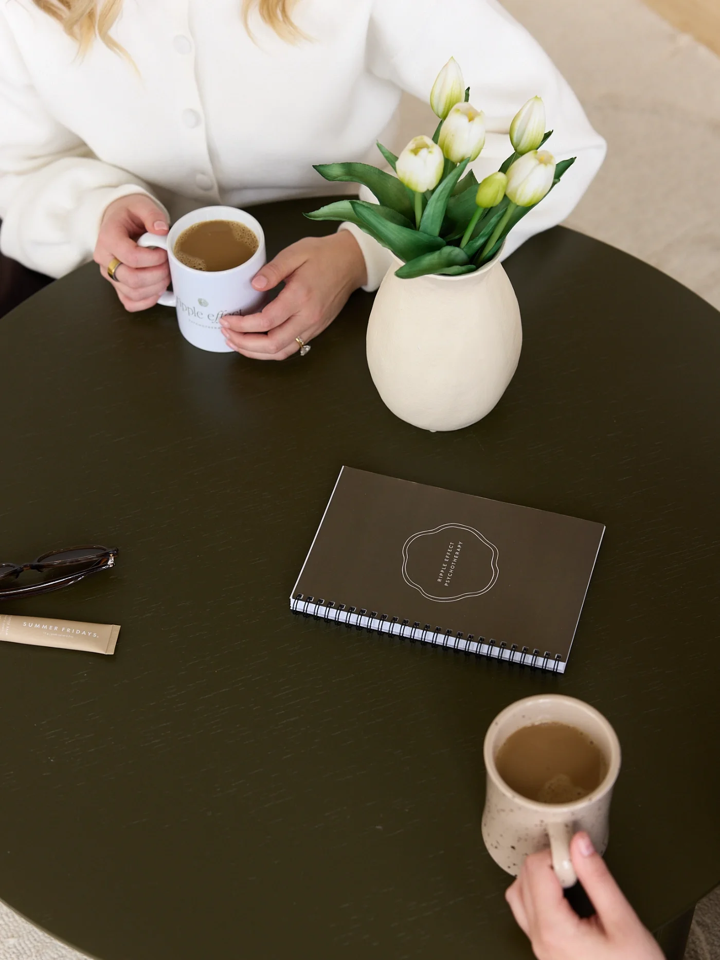 Two women holding coffees at a table, with a Ripple Effect Psychotherapy journal.