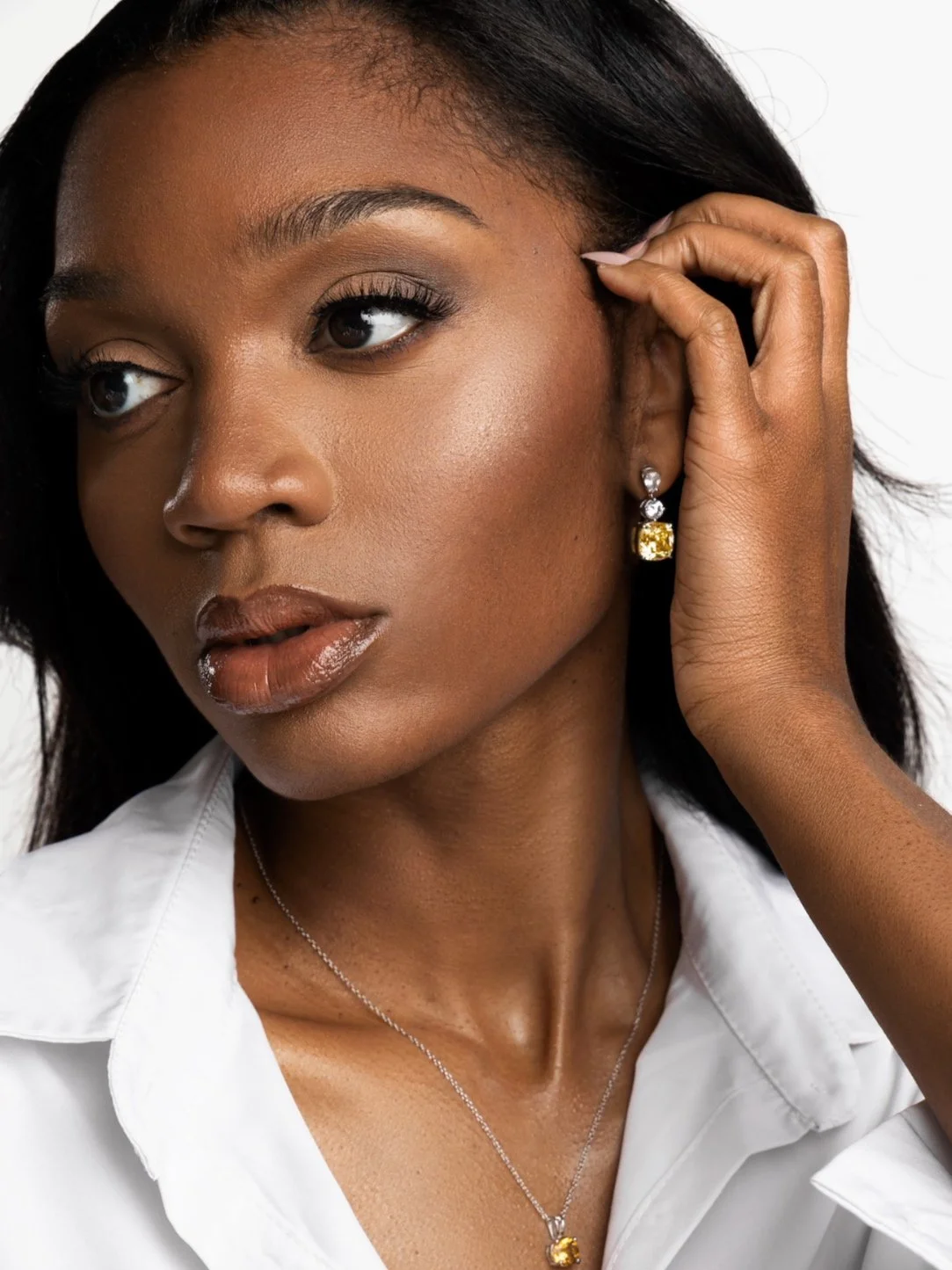 Close-up of a woman with dark skin, black hair, wearing a white shirt, gold and diamond earrings, and a matching necklace, touching her ear.