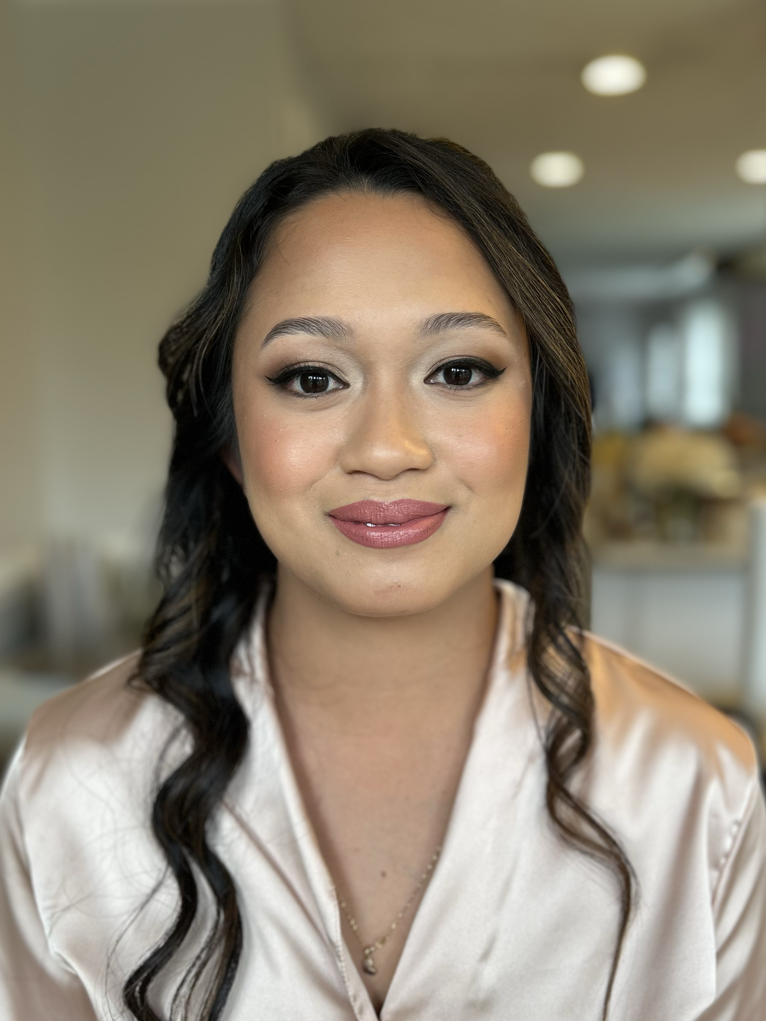 Close-up of a woman with styled black hair, wearing makeup with winged eyeliner and lipstick, smiling slightly at the camera, in an indoor setting.