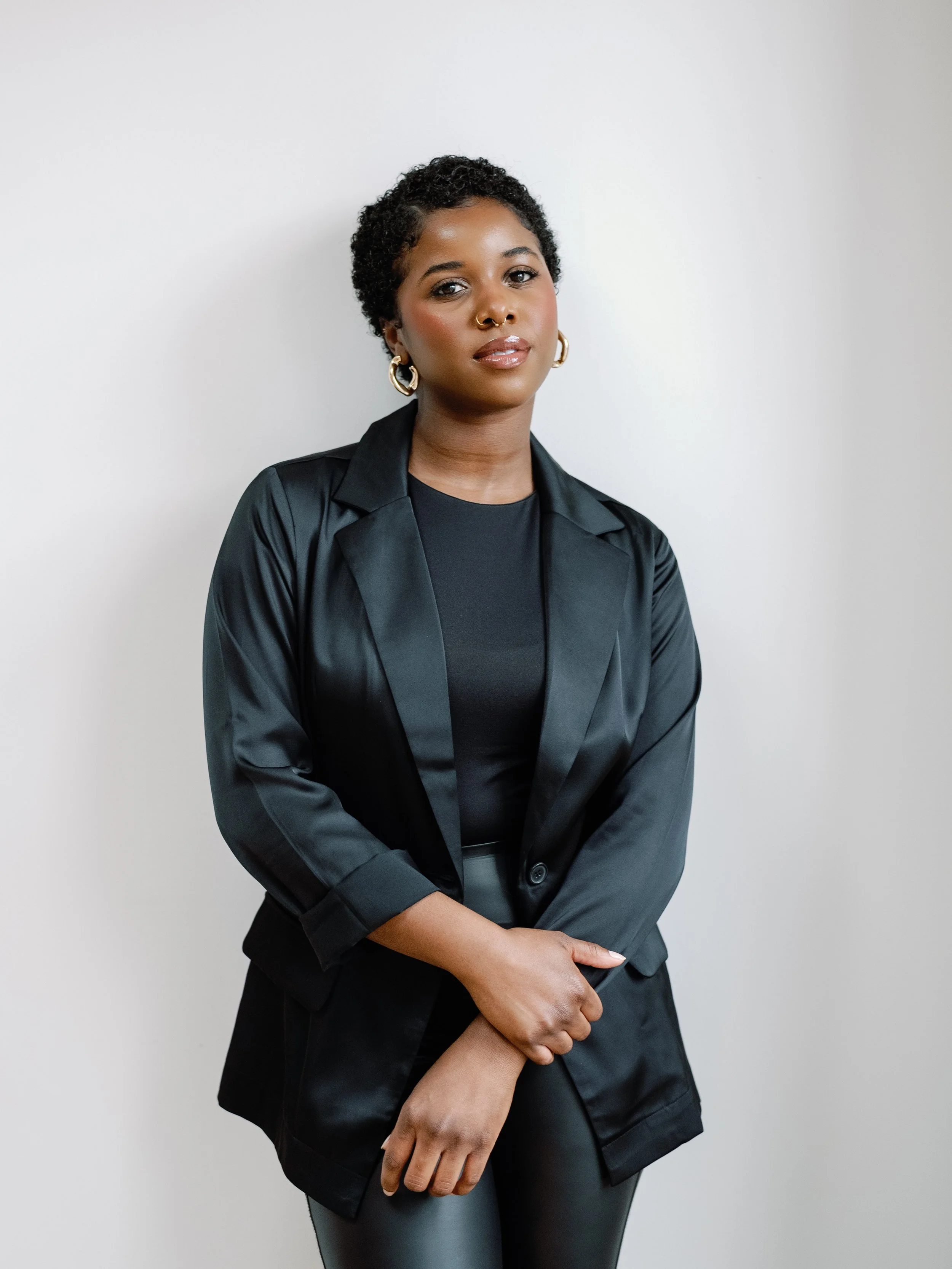 Portrait of a woman with short curly hair, wearing a black leather blazer, gold hoop earrings, and a septum piercing, standing against a plain white background.