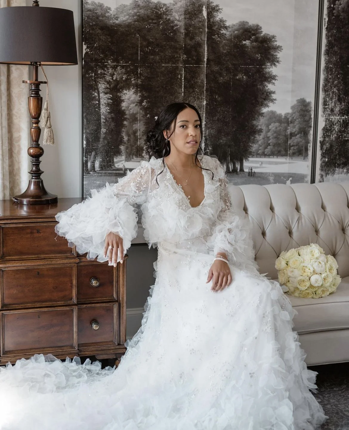 A woman in a white wedding gown with ruffled sleeves sitting on a cream-colored tufted sofa, with a bouquet of white roses beside her, standing beside a wooden side table and vintage-style lamp in a room with a large black-and-white landscape photogr