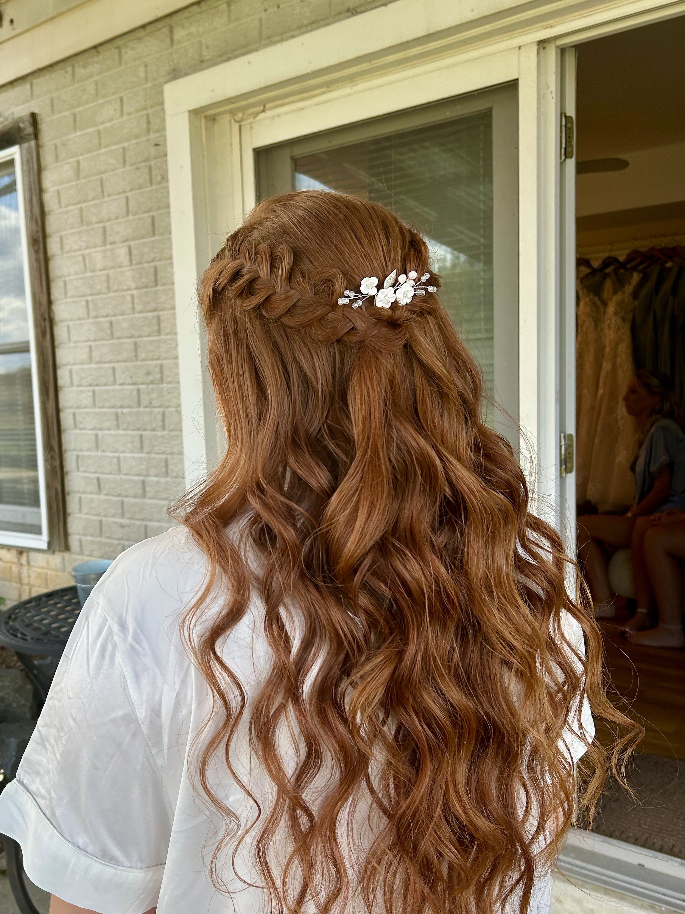 A woman with long, wavy red hair styled in a braid with a decorative white floral hairpin, standing outside near a window, with a person sitting inside visible in the background.