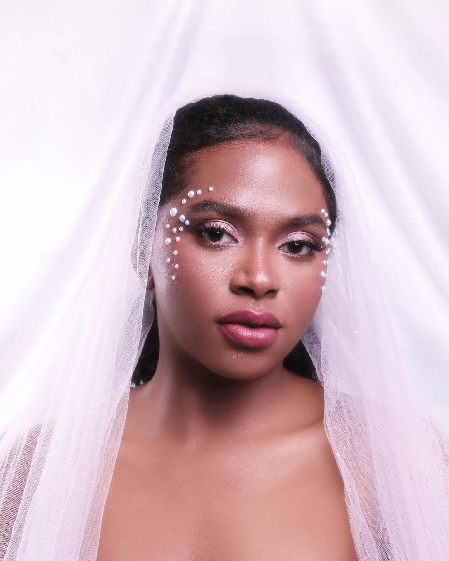 Close-up of a woman with dark hair styled in a sleek updo, wearing a white bridal veil and pearl embellishments around her eyes, with makeup featuring pink eyeshadow and lipstick, posing against a white background.