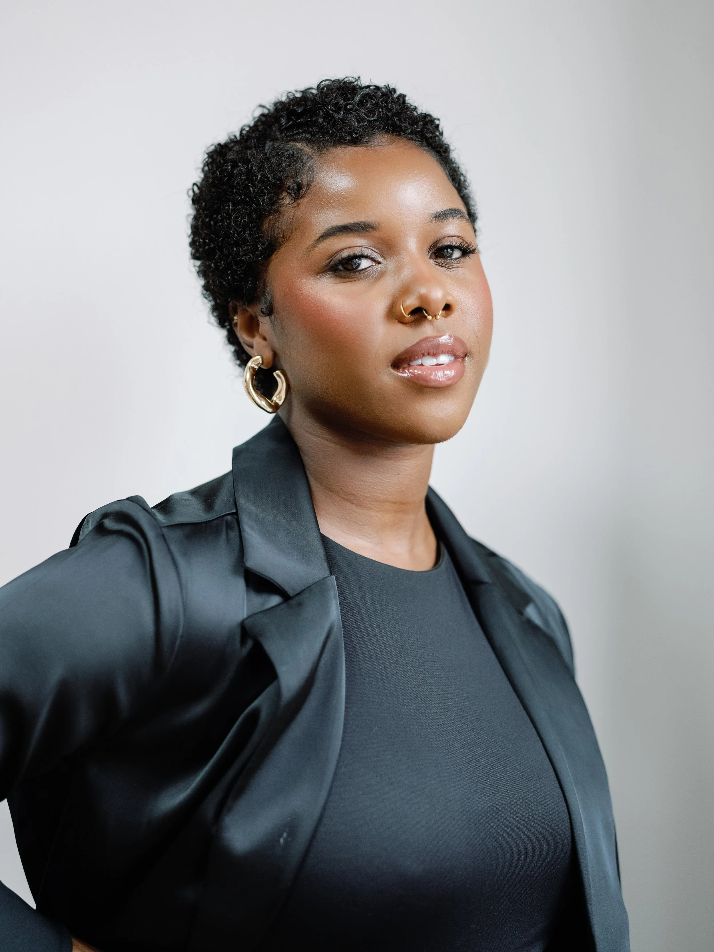 Portrait of a woman with short, curly black hair, dark skin, wearing gold hoop earrings, a nose ring, and a black top. She is looking at the camera with a confident expression against a plain light gray background.