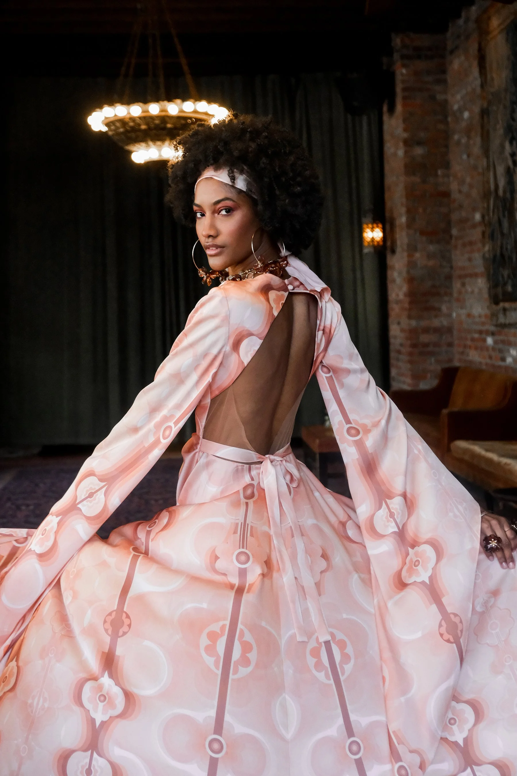 A woman with curly hair wearing a pink patterned dress with open back, sitting in a dimly lit room with brick and wood walls and a large chandelier hanging from the ceiling.