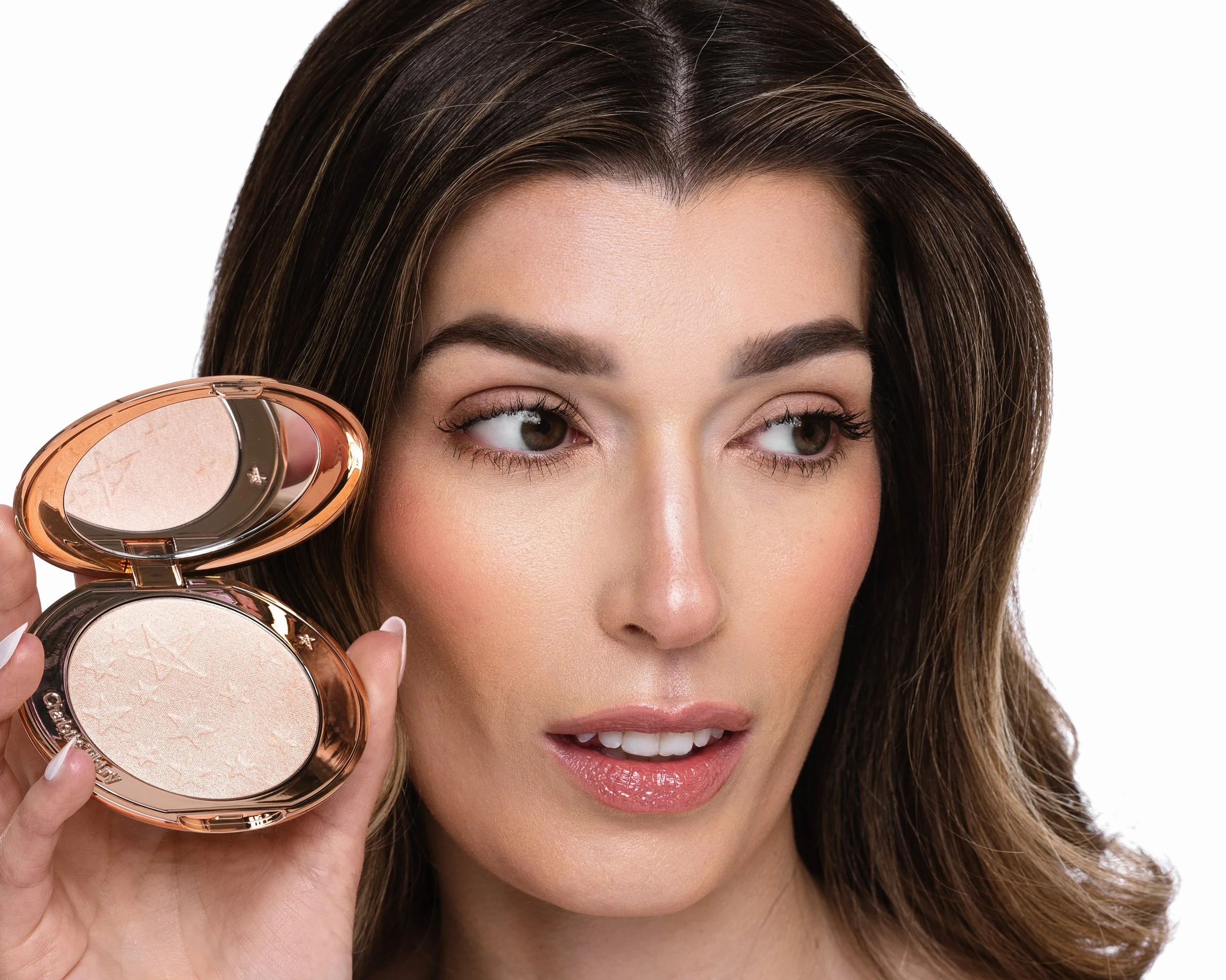 A woman with brown hair styled straight and parted in the middle, holding a compact powder case near her face, and smiling slightly with pink lips, against a plain white background.