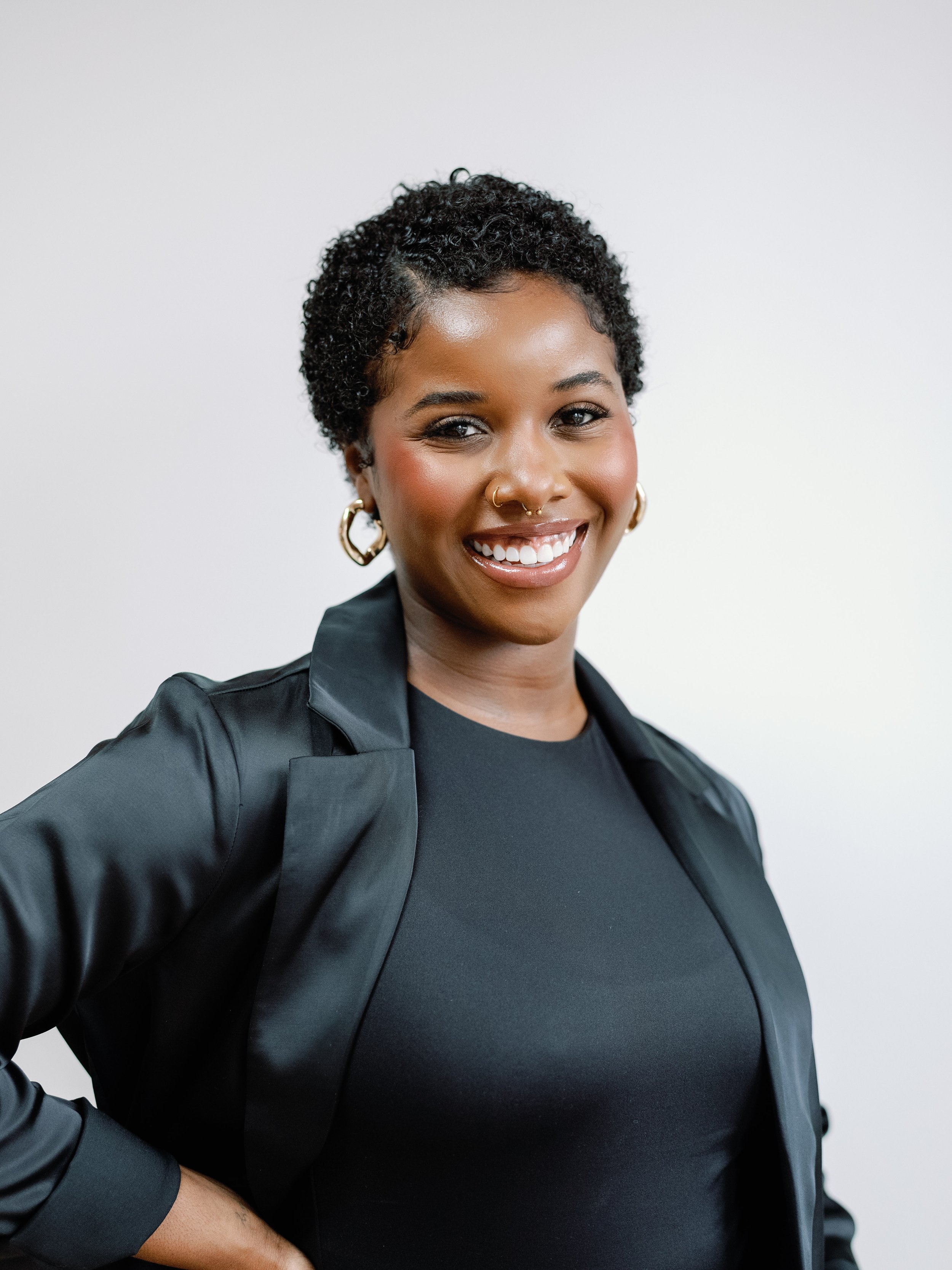 A young woman with short, curly black hair and brown skin, smiling and looking at the camera. She is wearing gold hoop earrings, a piercings on her nose and septum, and a black blazer over a black top, standing against a plain light background.