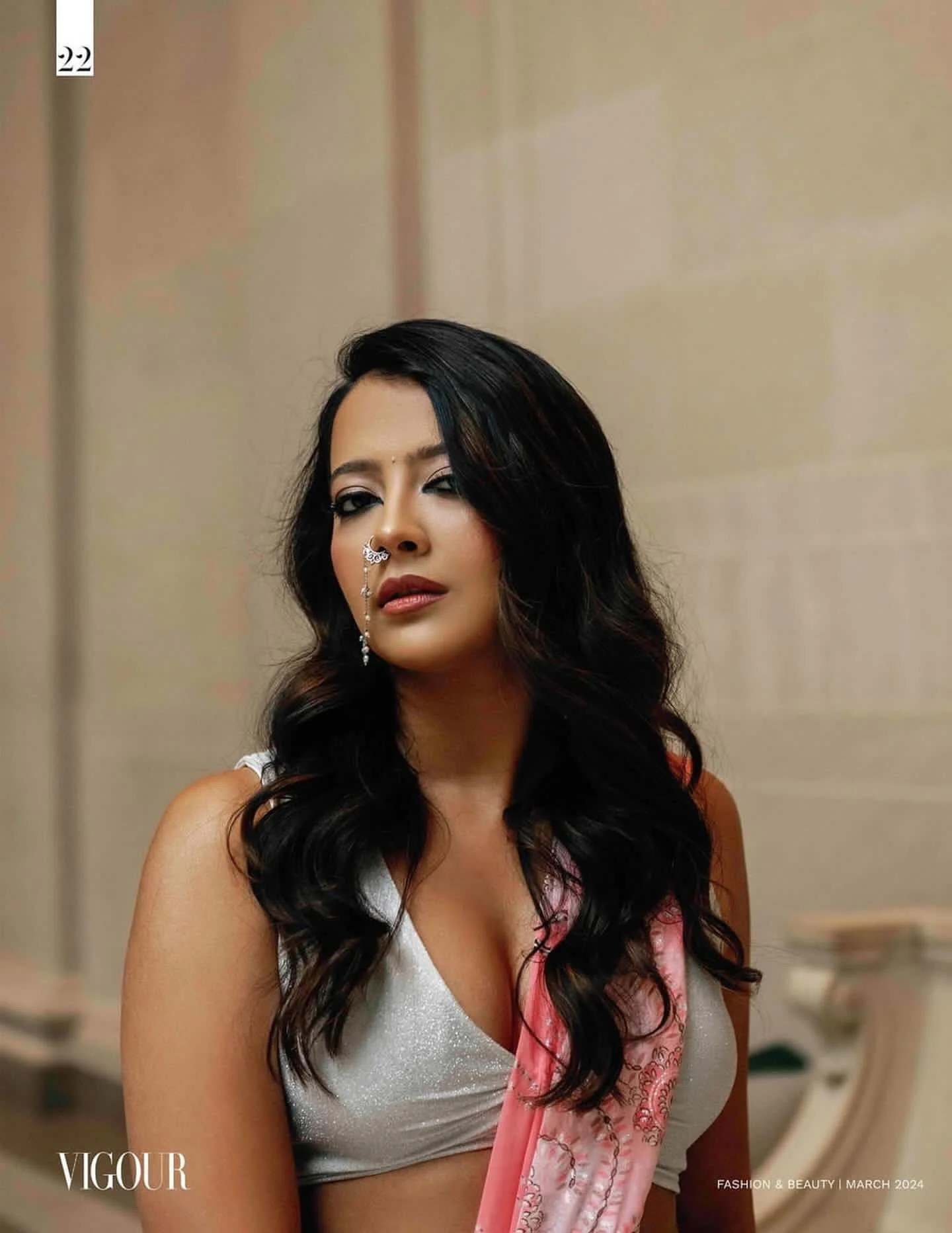 A woman with long black wavy hair, wearing a white top with a pink floral scarf, standing against a beige background with soft lighting.