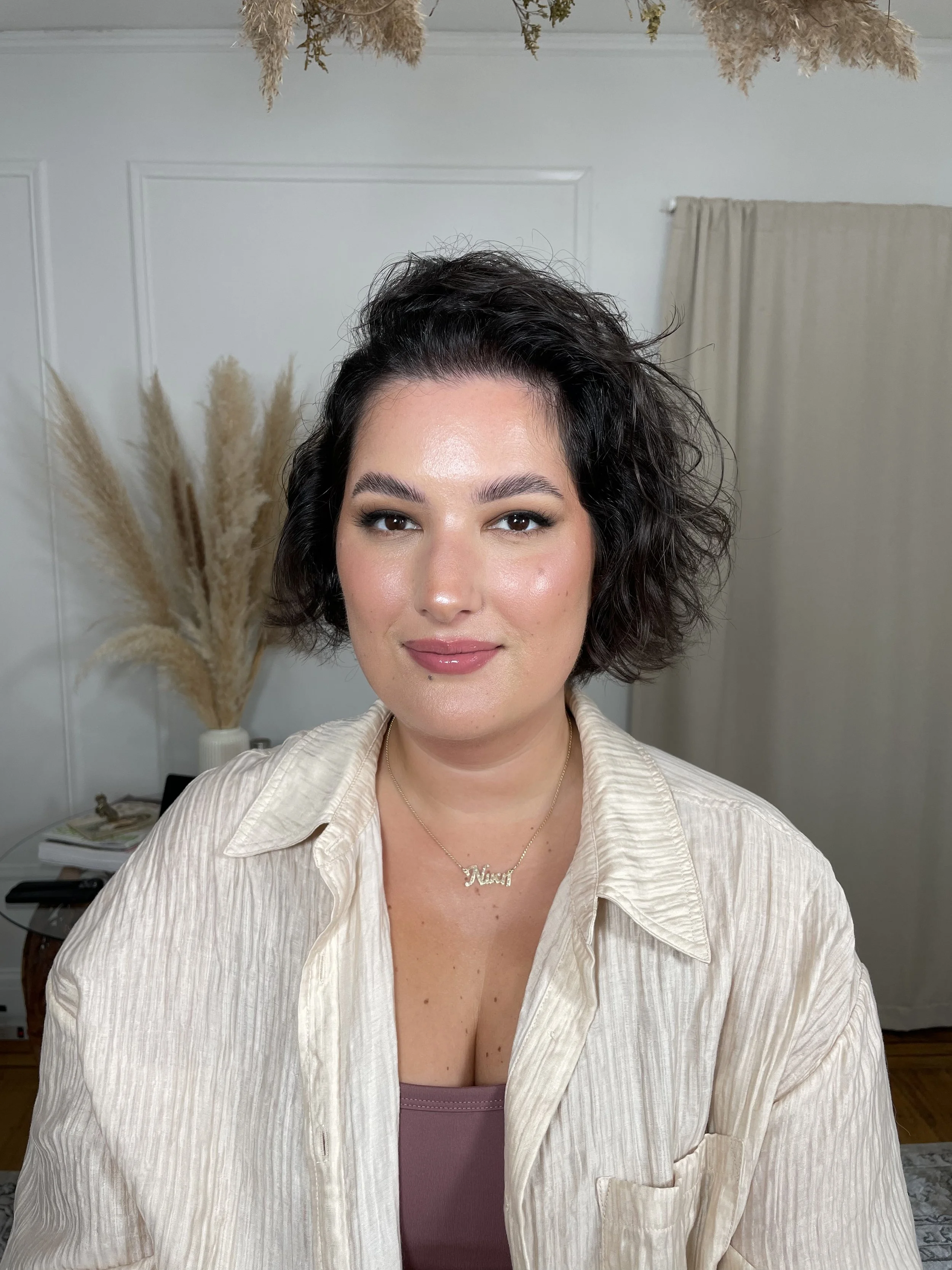 A woman with dark, curly, short hair smiling at the camera. She is wearing a beige, striped shirt over a mauve top, accessorized with a necklace that spells "Nicki." The background includes pampas grass in a white vase, a closed beige curtain, and a 