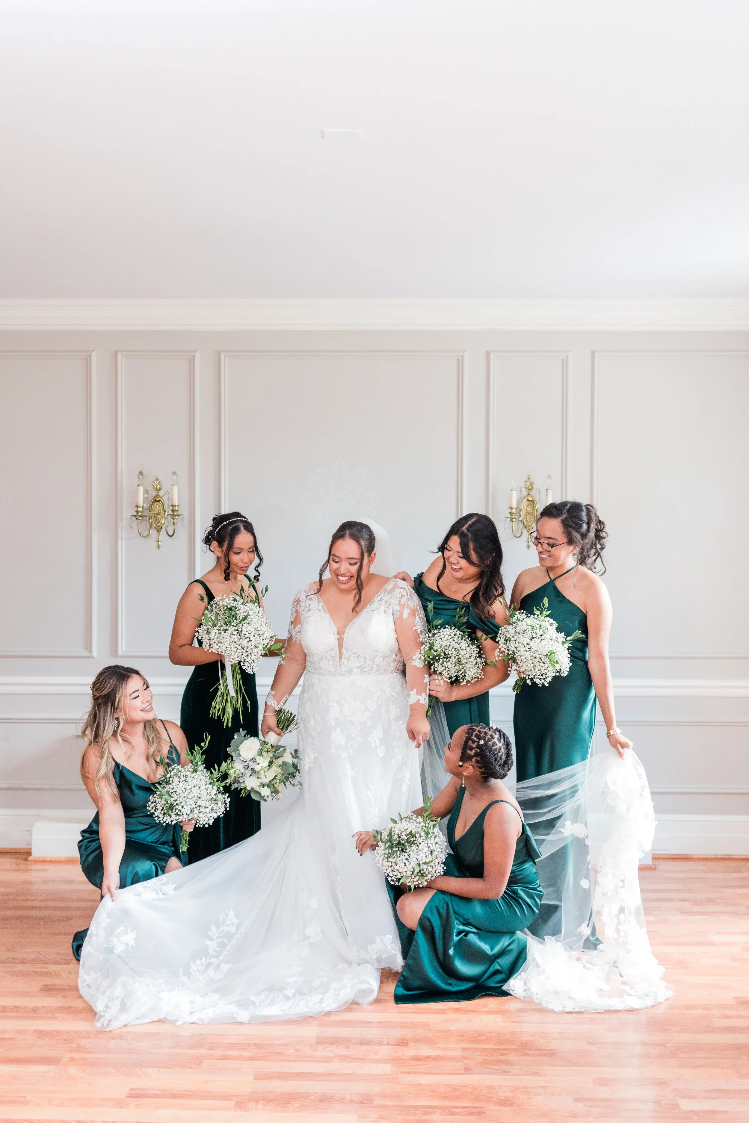 A wedding ceremony taking place indoors with a white wall and wooden floor. The bride is in a white lace wedding dress, surrounded by bridesmaids in dark teal dresses holding bouquets of white flowers. The bride is smiling and the bridesmaids are ass