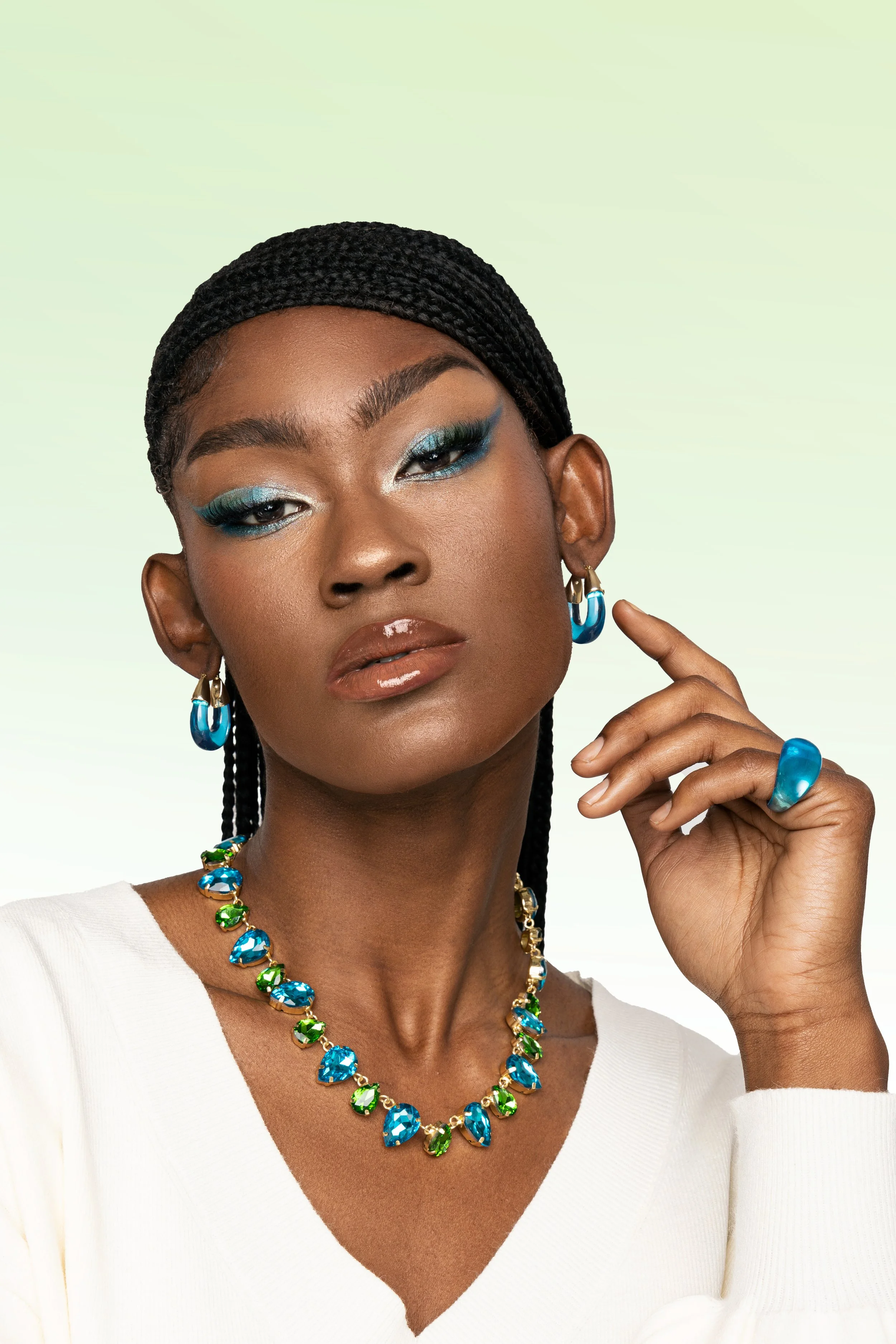 A woman with dreadlocks wearing blue and green jewelry, makeup, and a white top against a light green background.