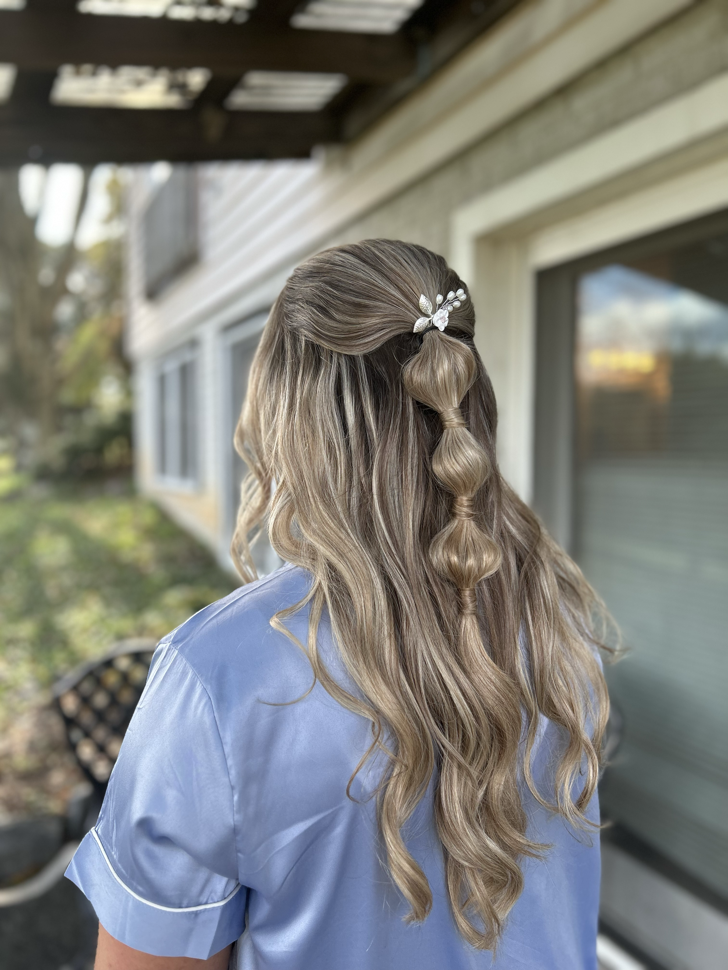 Back view of a woman with long wavy blonde hair styled with a decorative hair clip, wearing a light blue blouse, standing outdoors near a house with white siding and windows.