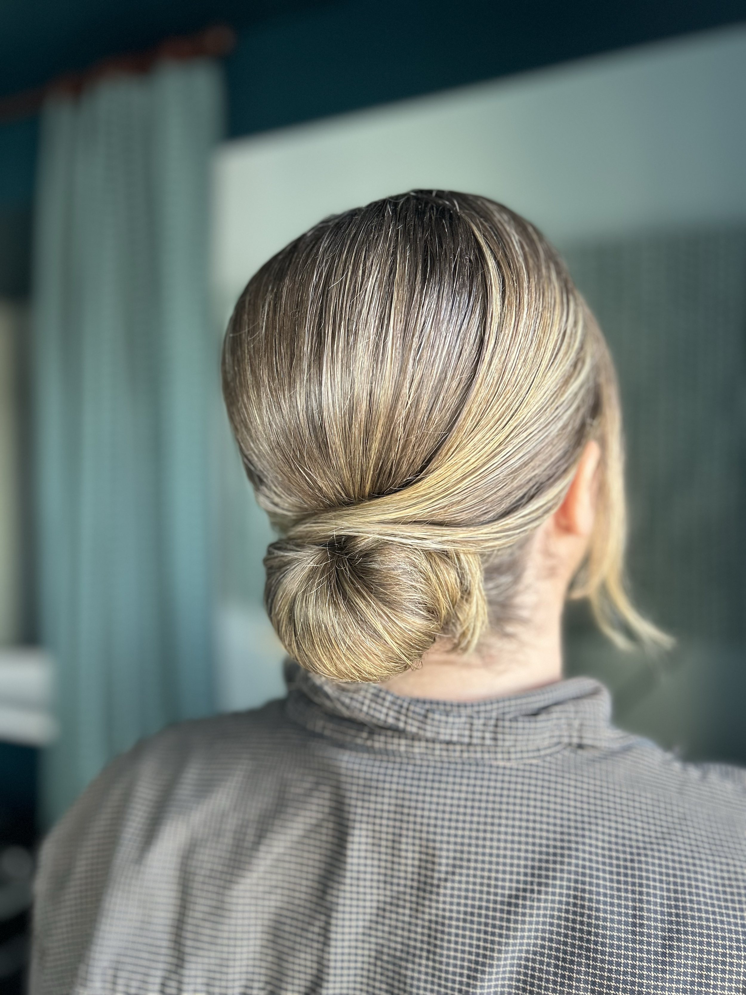 Back of a woman's head with a styled low bun hairstyle, blonde hair with darker roots, wearing a checkered shirt, in front of a blurred background with teal and beige curtains.