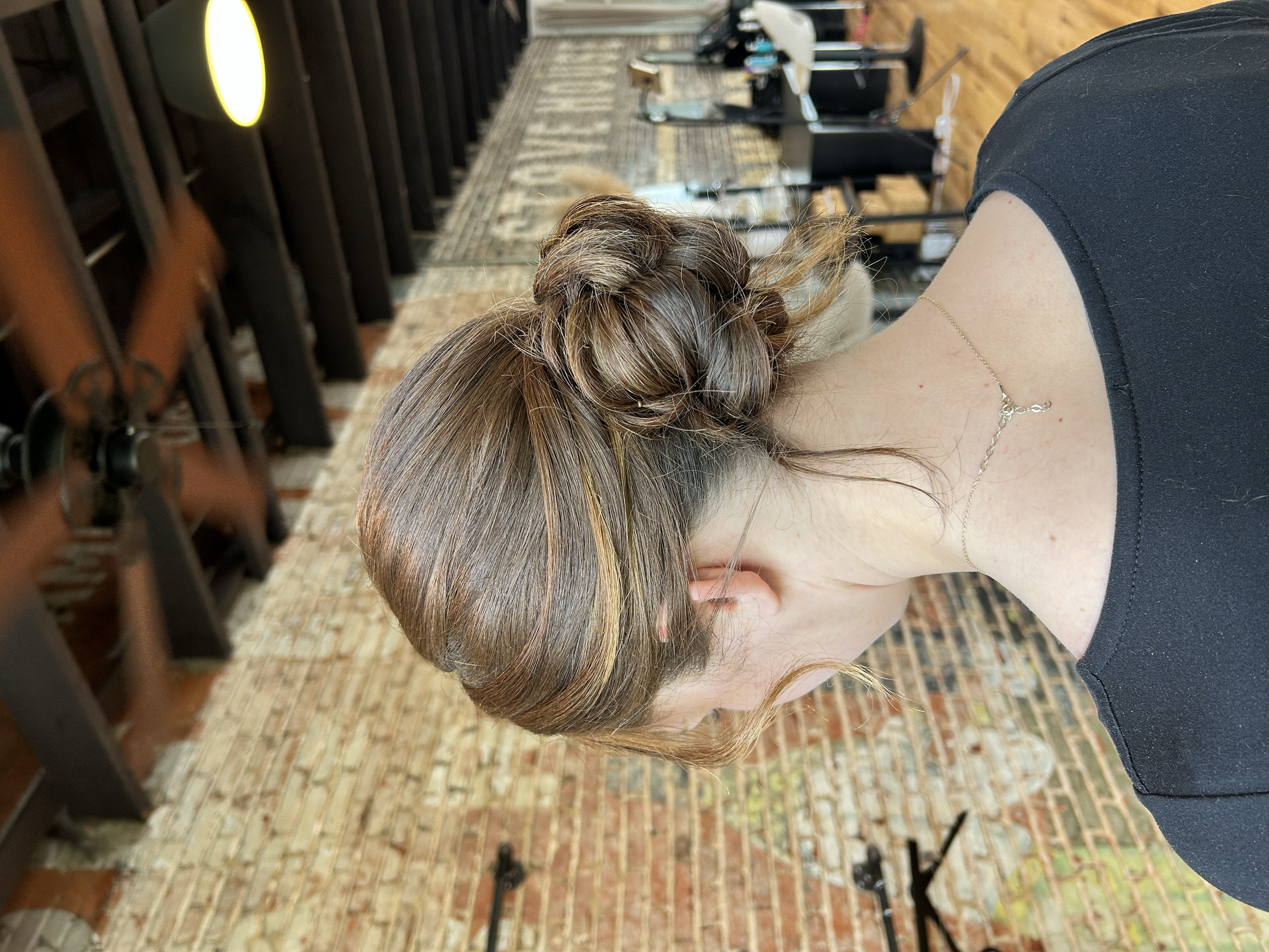 The image shows a person with brown hair styled in an elegant bun, wearing a black top and a delicate silver necklace, sitting in a salon with a brick floor and wooden wall decor, and hair styling equipment visible in the background.