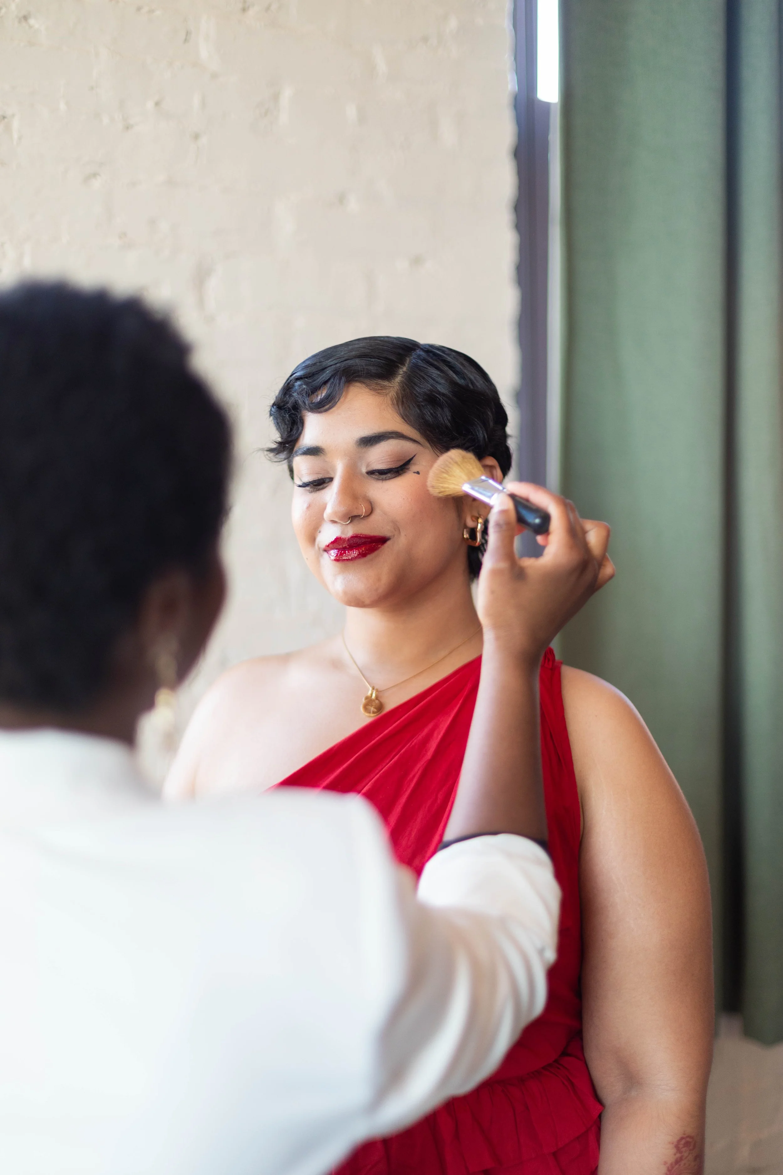 A woman with short black hair and red lipstick is having her makeup done with a brush by a person whose face is not visible. She is wearing a red dress and a gold necklace, smiling with closed eyes, standing near a green curtain and a beige wall.