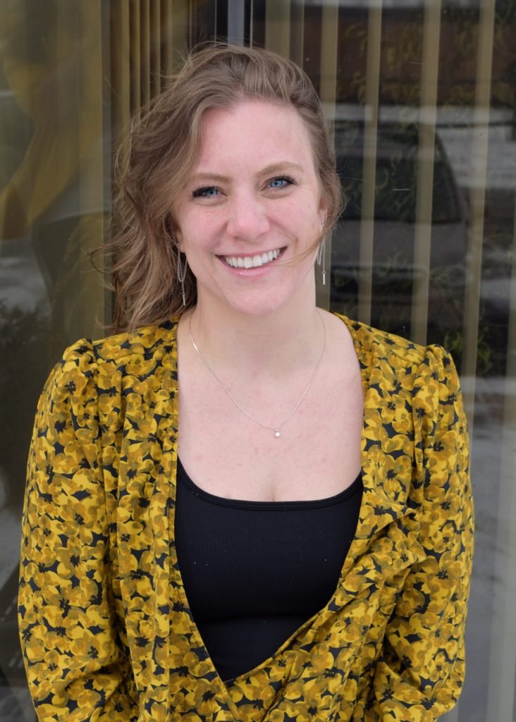 A woman with wavy light brown hair, blue eyes, and fair skin is smiling outdoors. She is wearing a black top and a yellow floral-patterned blazer. She has delicate earrings and a small necklace, standing in front of a glass building with reflections.