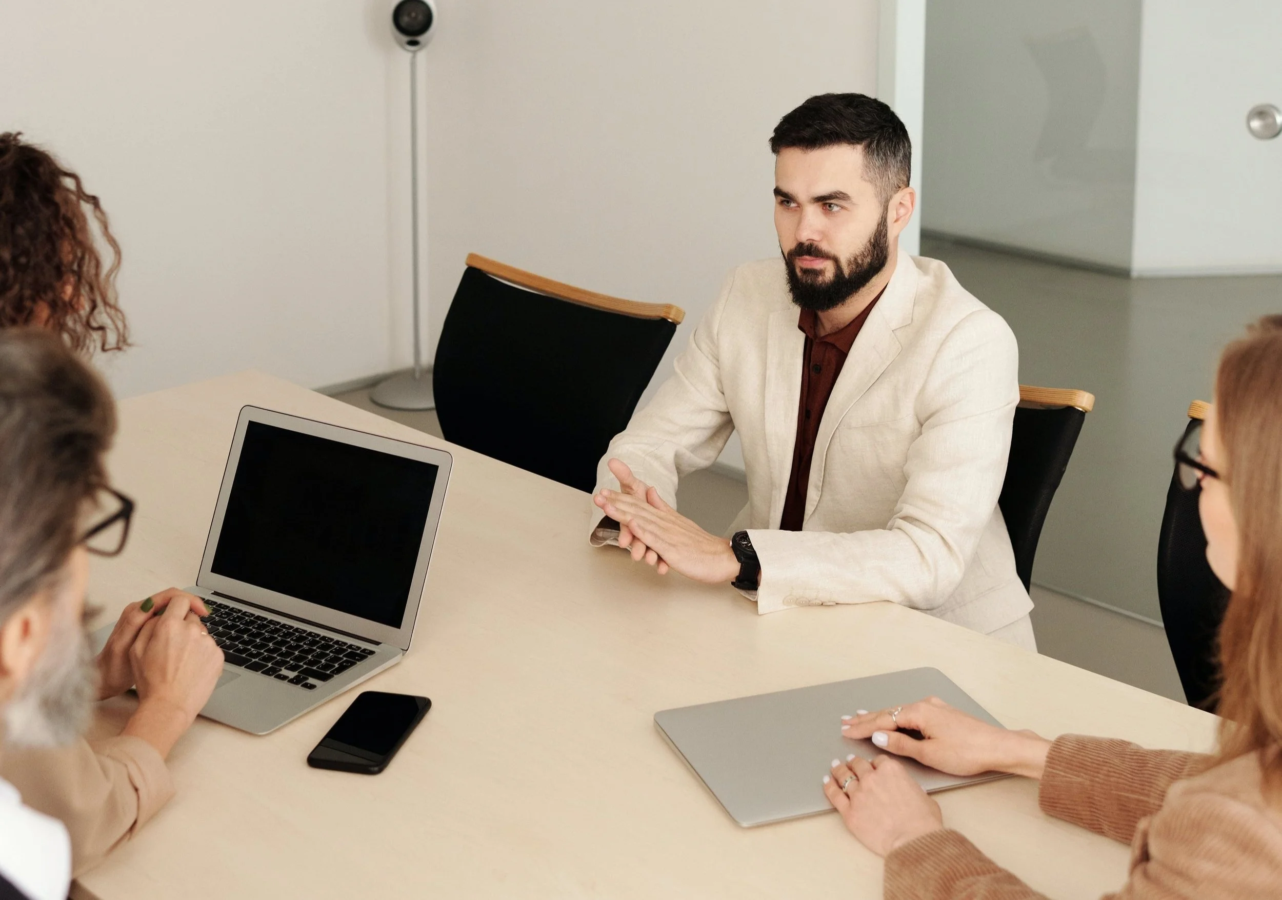 A man with a beard, wearing a beige blazer, is sitting at a conference table with three women. They are in a meeting room, engaging in a discussion. There is a laptop, a smartphone, and a closed notebook on the table.
