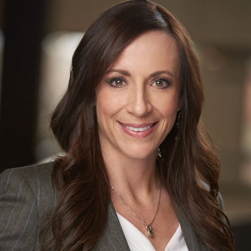 A woman with long brown hair wearing a gray blazer and white top, smiling and looking directly at the camera.